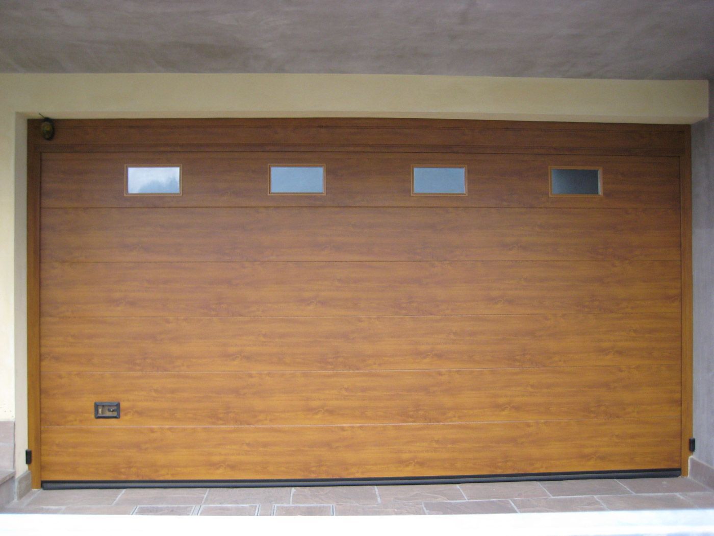Porta da garage in legno con quattro piccole finestre, incastonata in un muro esterno in cemento.