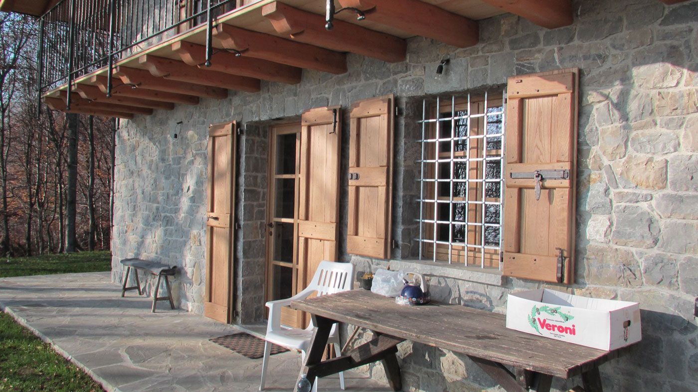 Esterno rustico di una casa in pietra con persiane in legno, tavolo da patio e panchina sotto un balcone.