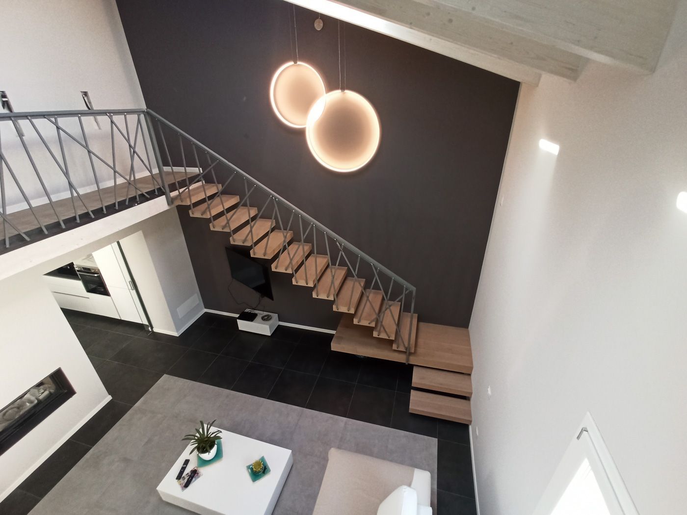 Soggiorno moderno con scala sospesa, parete d'accento nera e lampade a sospensione sopra una zona salotto.