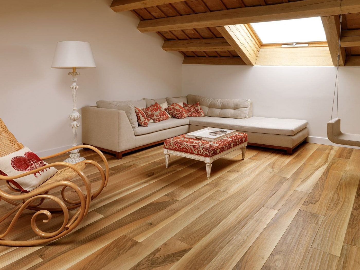 Accogliente soggiorno mansardato con divano angolare beige, poltrona in vimini, pouf a fantasia e soffitto in legno inclinato.