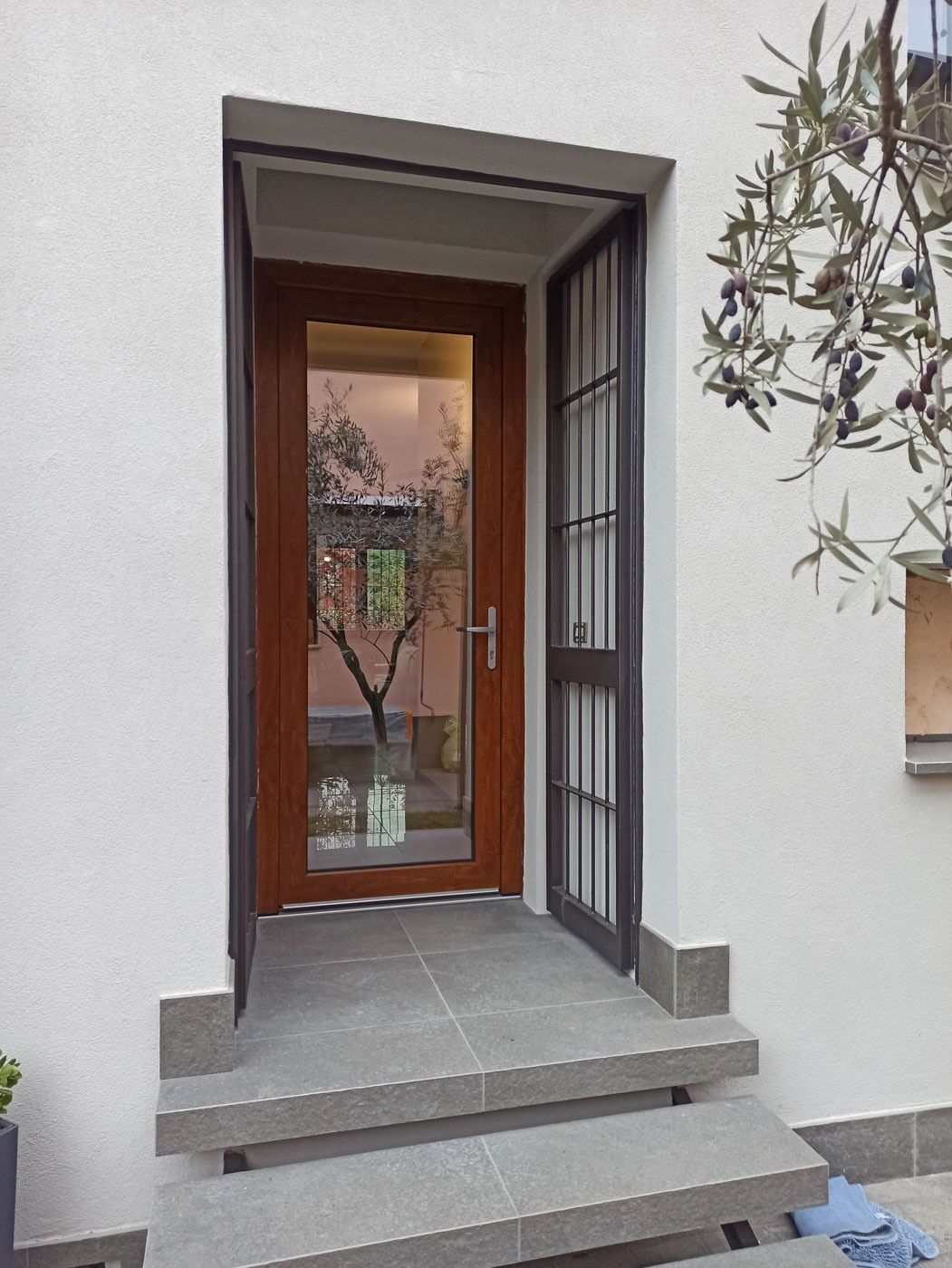 Ingresso principale con scalini, muri in stucco bianco, porta in legno e cancello di sicurezza in metallo che si apre su un cortile.