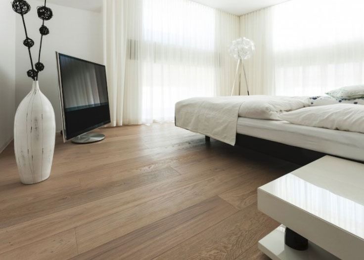 Camera da letto minimalista con letto bianco, pavimento in legno, ampie finestre e televisore inclinato sul pavimento.