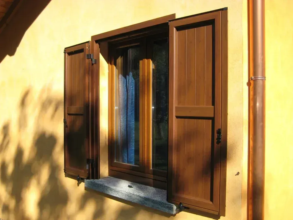 Persiane di legno aperte su una parete di stucco giallo, con giochi di luce e ombre.
