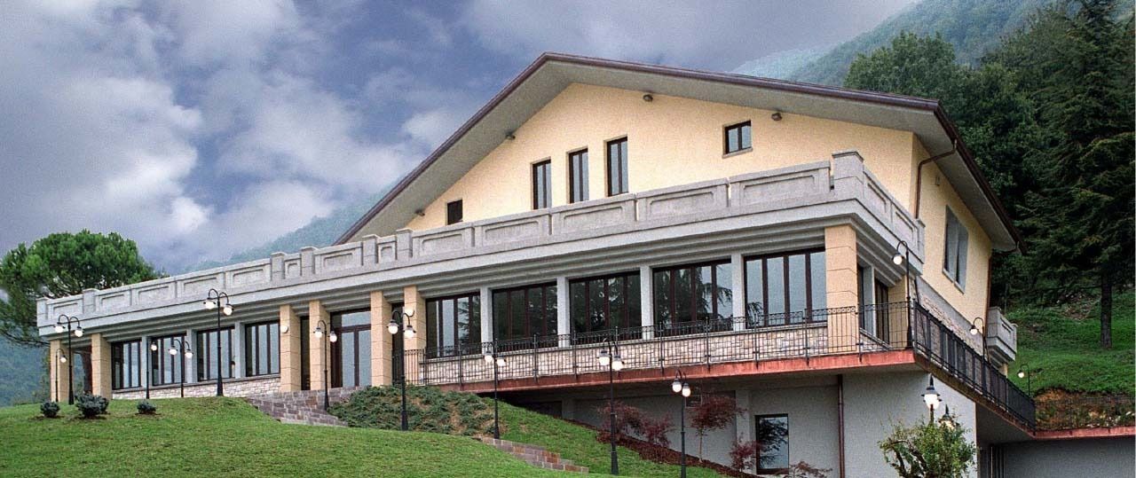 Rifugio di montagna con lungo balcone e grandi finestre su un pendio erboso sotto un cielo nuvoloso.