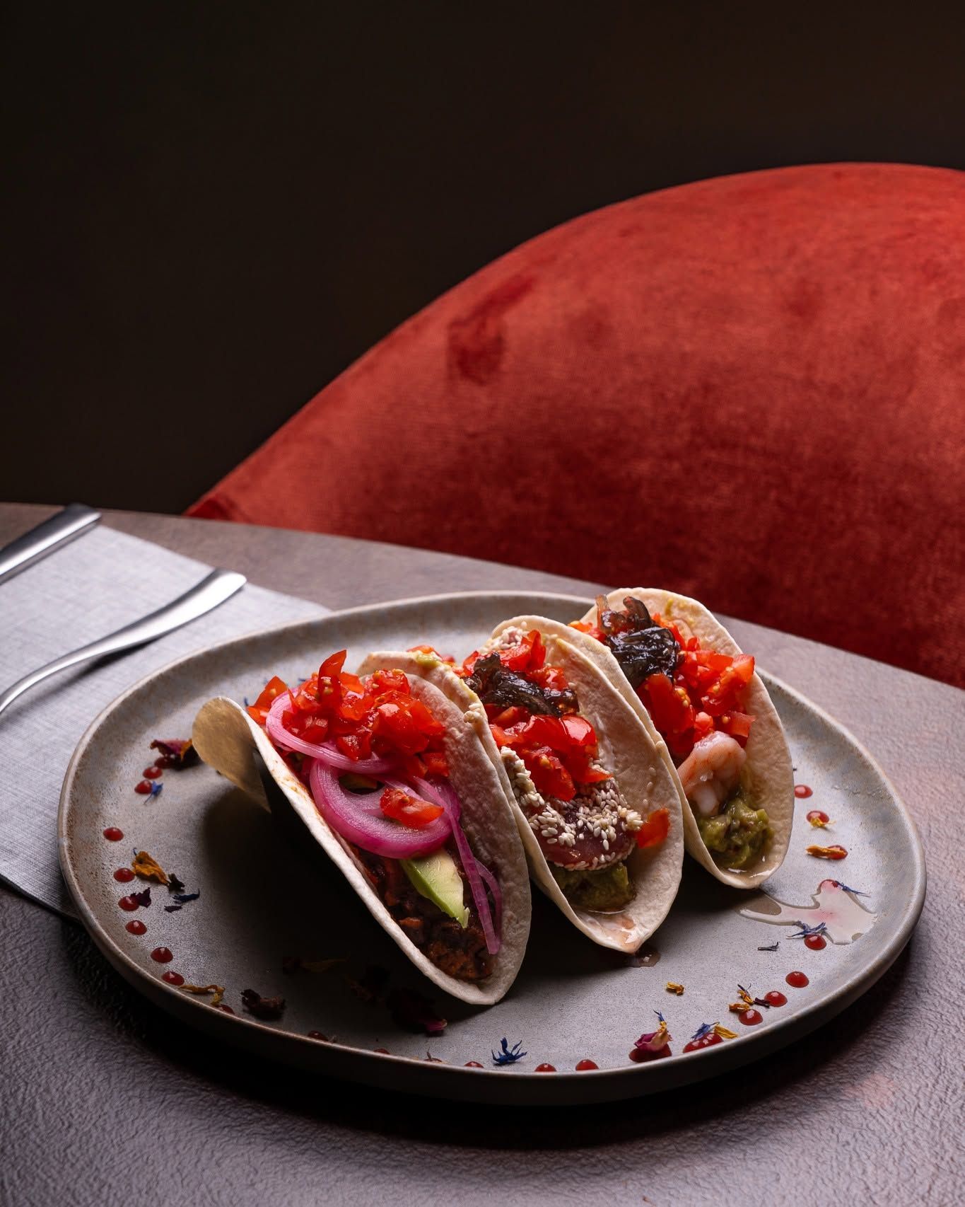 Tre tacos ben farciti su un piatto grigio accanto a una forchetta su un tavolo scuro con una sedia rossa sullo sfondo.