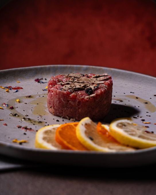 Medaglione di bistecca scottato su un piatto grigio con fette d'arancia e di limone, su uno sfondo rosso scuro.