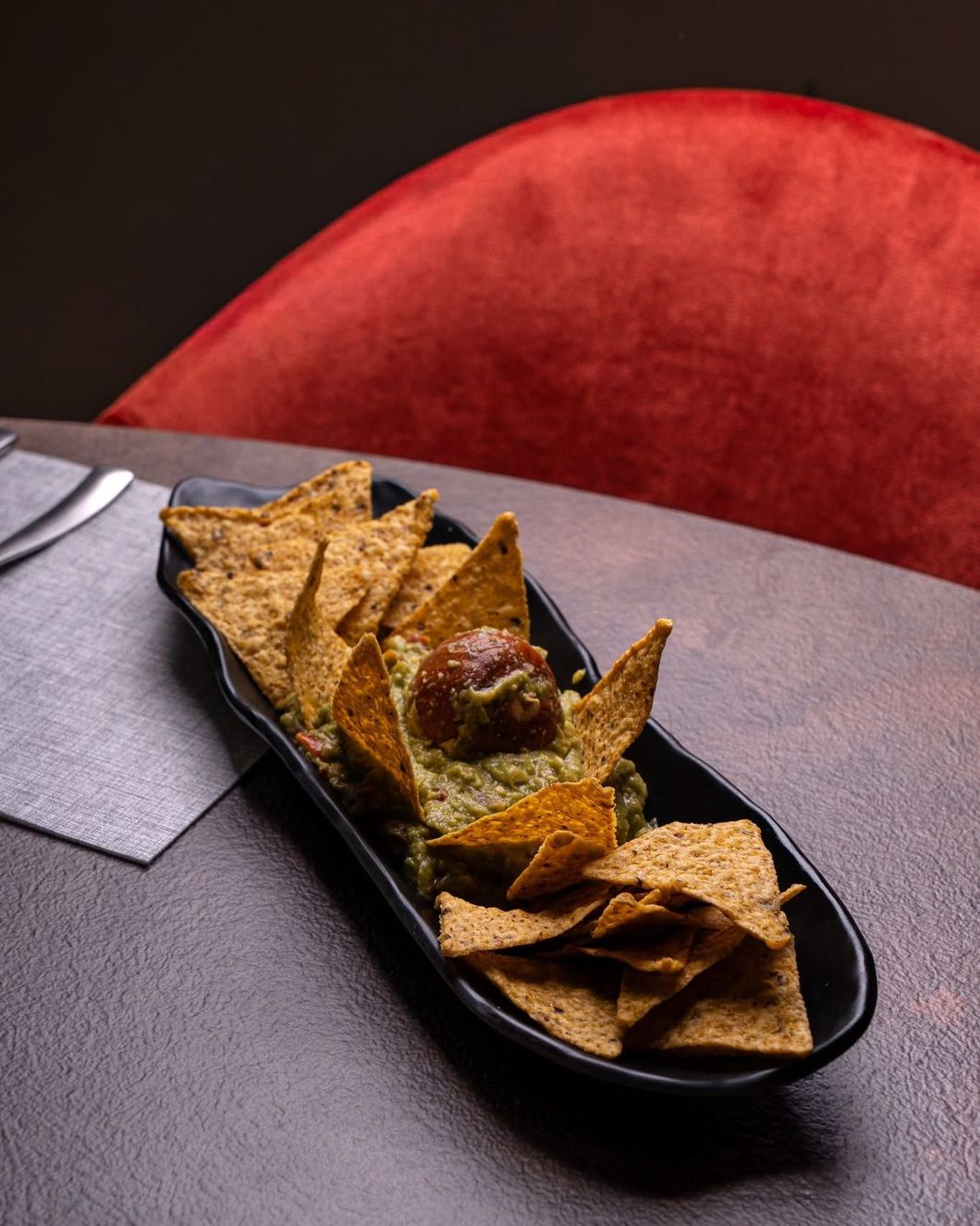 Tortilla chips con salsa in un piatto da portata nero su un tavolo, con una sedia imbottita rossa alle sue spalle.