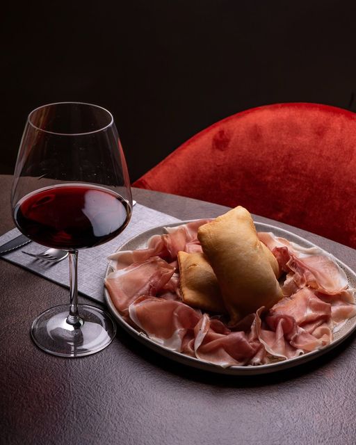 Un bicchiere di vino e un piatto di prosciutto con un panino su un tavolo, illuminati sullo sfondo di una sedia rossa.