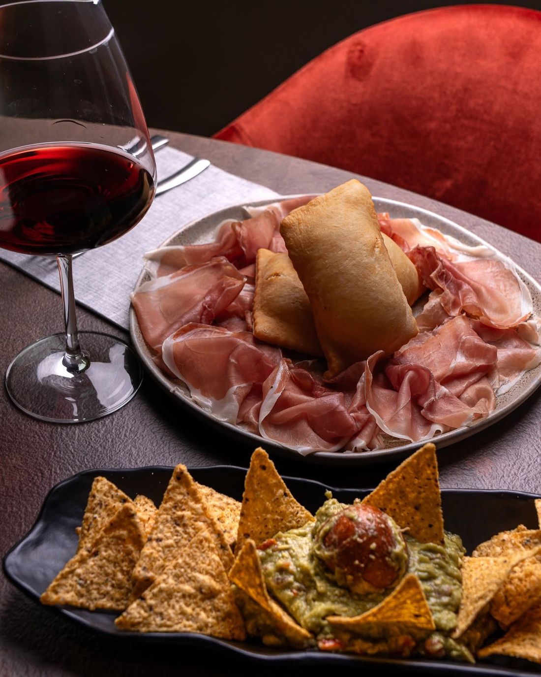 Un calice di vino accanto a un tagliere di salumi con prosciutto e un grissino, accompagnato da tortilla chips con salsa.