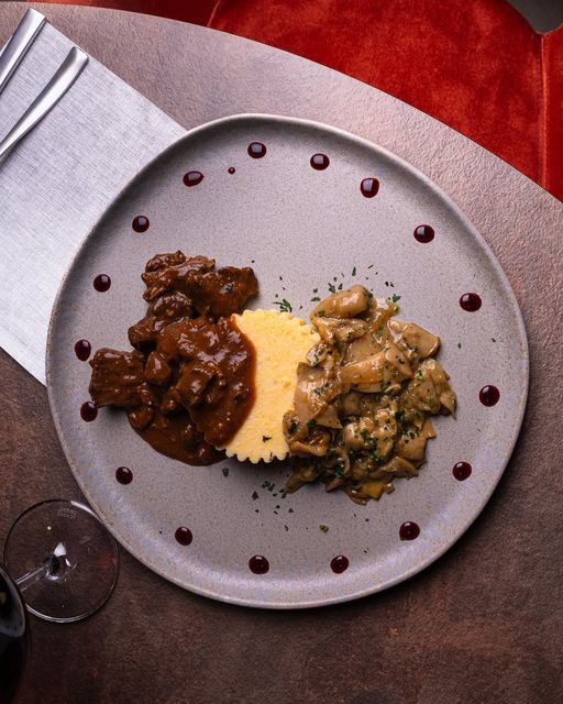 Due piatti con salsa su un piatto grigio con una fetta di pane, su un tavolo con calice da vino e posate.