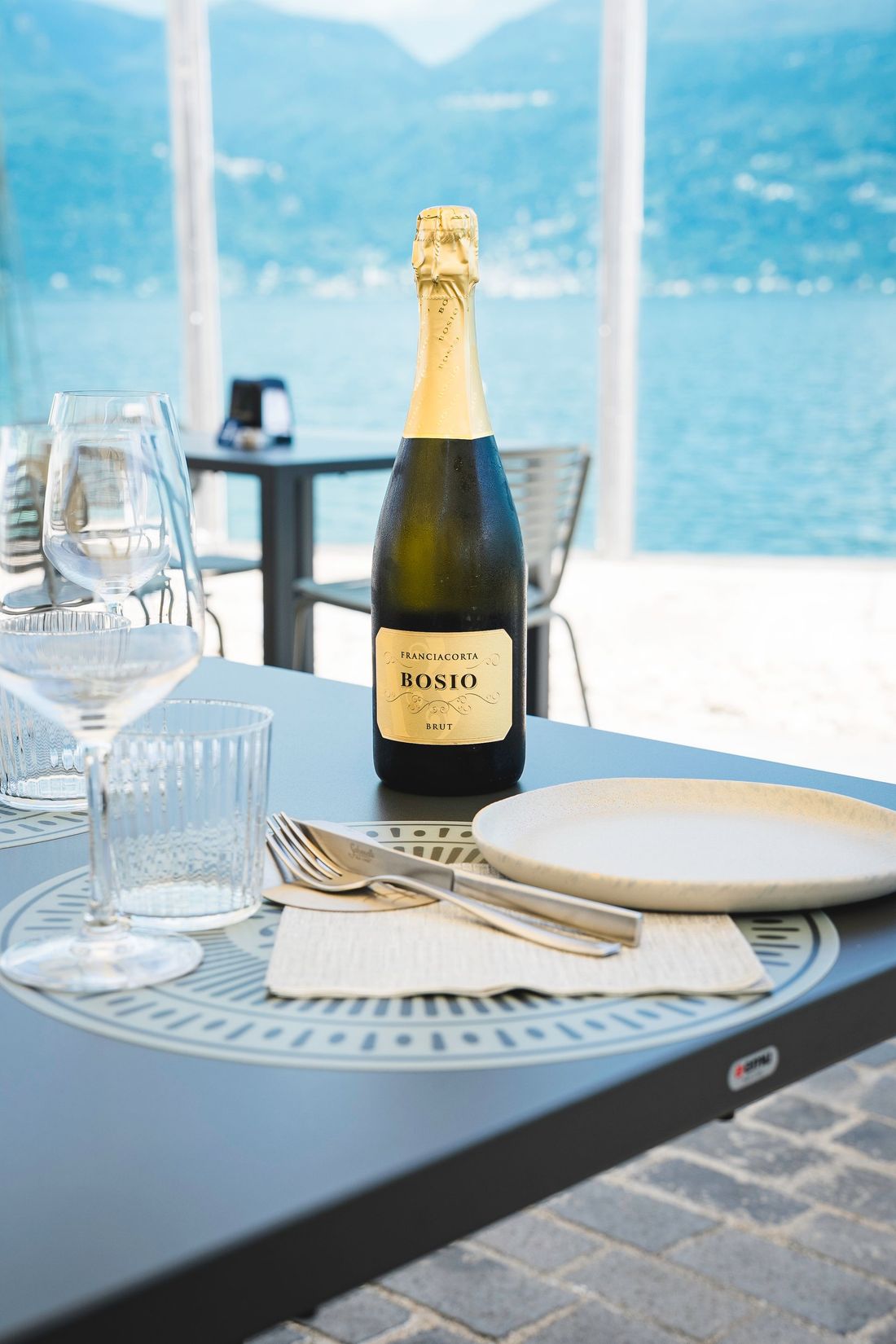 Una bottiglia di champagne su un tavolo in riva al mare con bicchieri e piatti, con vista sull'acqua azzurra e sulle montagne.