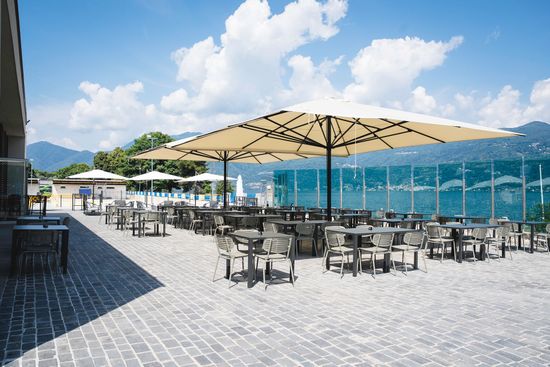 Terrazza esterna del caffè con tavolini, grandi ombrelloni beige e vista sulle montagne sotto un cielo azzurro brillante.
