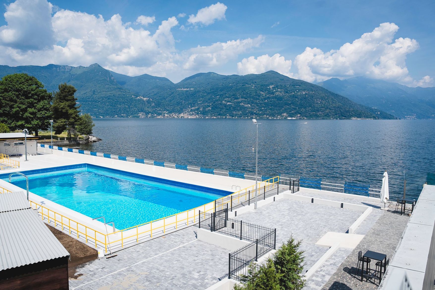 Terrazza a bordo lago con piscina, acque turchesi, lettini bianchi e vista sulle montagne sotto un cielo azzurro.