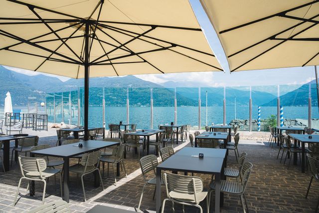 Terrazza esterna del ristorante con tavoli e ombrelloni vuoti, affacciata su un mare blu e sulle montagne.