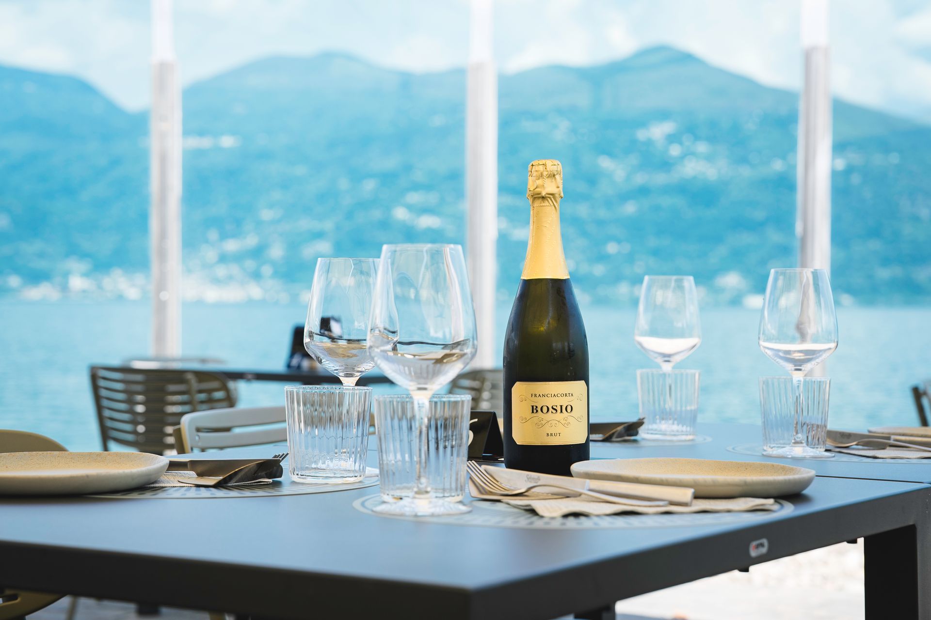 Tavolo da pranzo apparecchiato all'aperto con bicchieri da vino e bottiglia di champagne, con vista su un lago e sulle montagne.