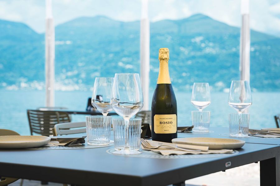 Tavolo da pranzo all'aperto con bicchieri da vino e bottiglia di champagne, con vista su un lago e sulle montagne.