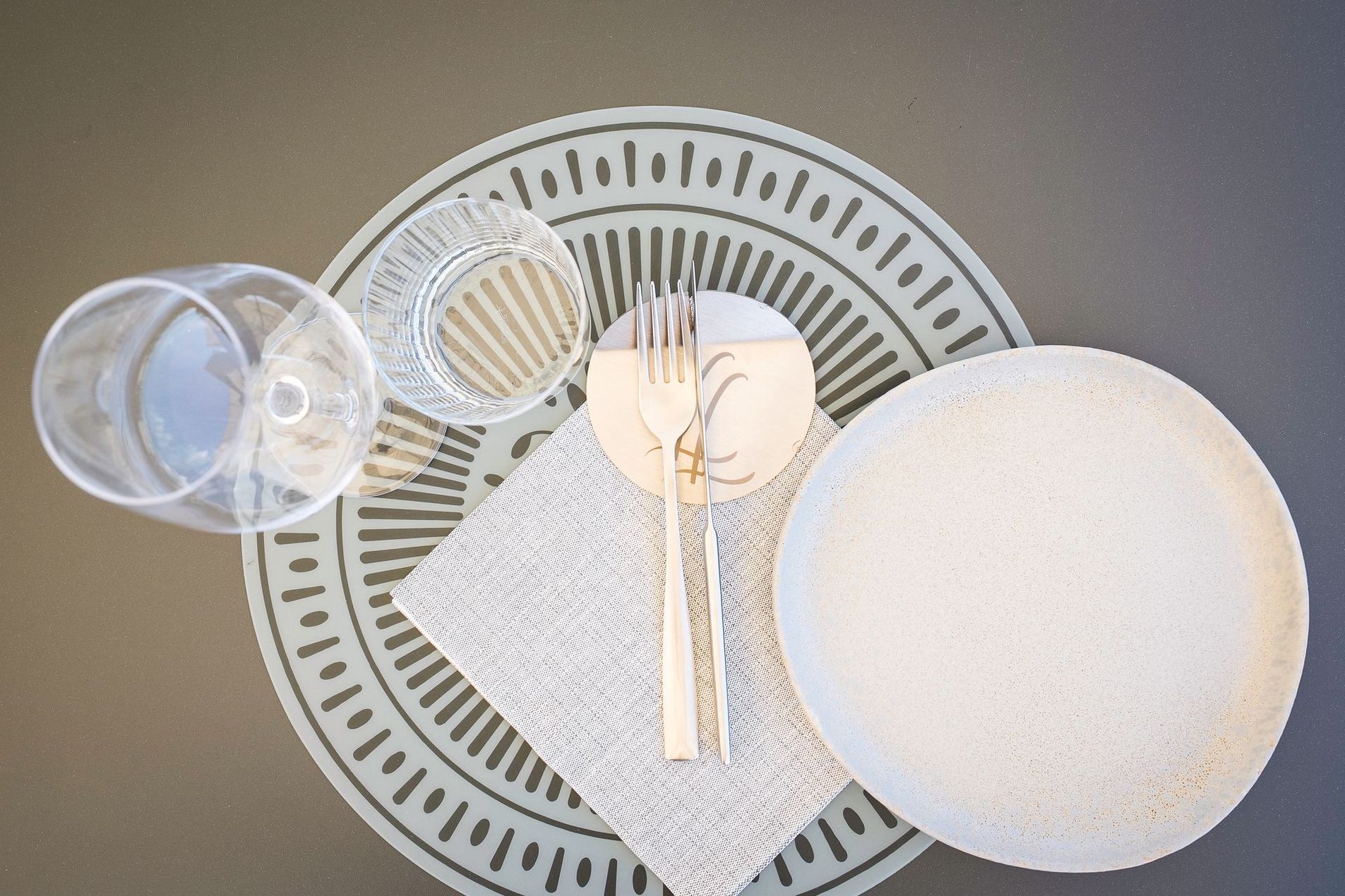 Tavola apparecchiata con piatti bianchi, bicchieri trasparenti, posate dorate e un tovagliolo su un sottopiatto decorato.