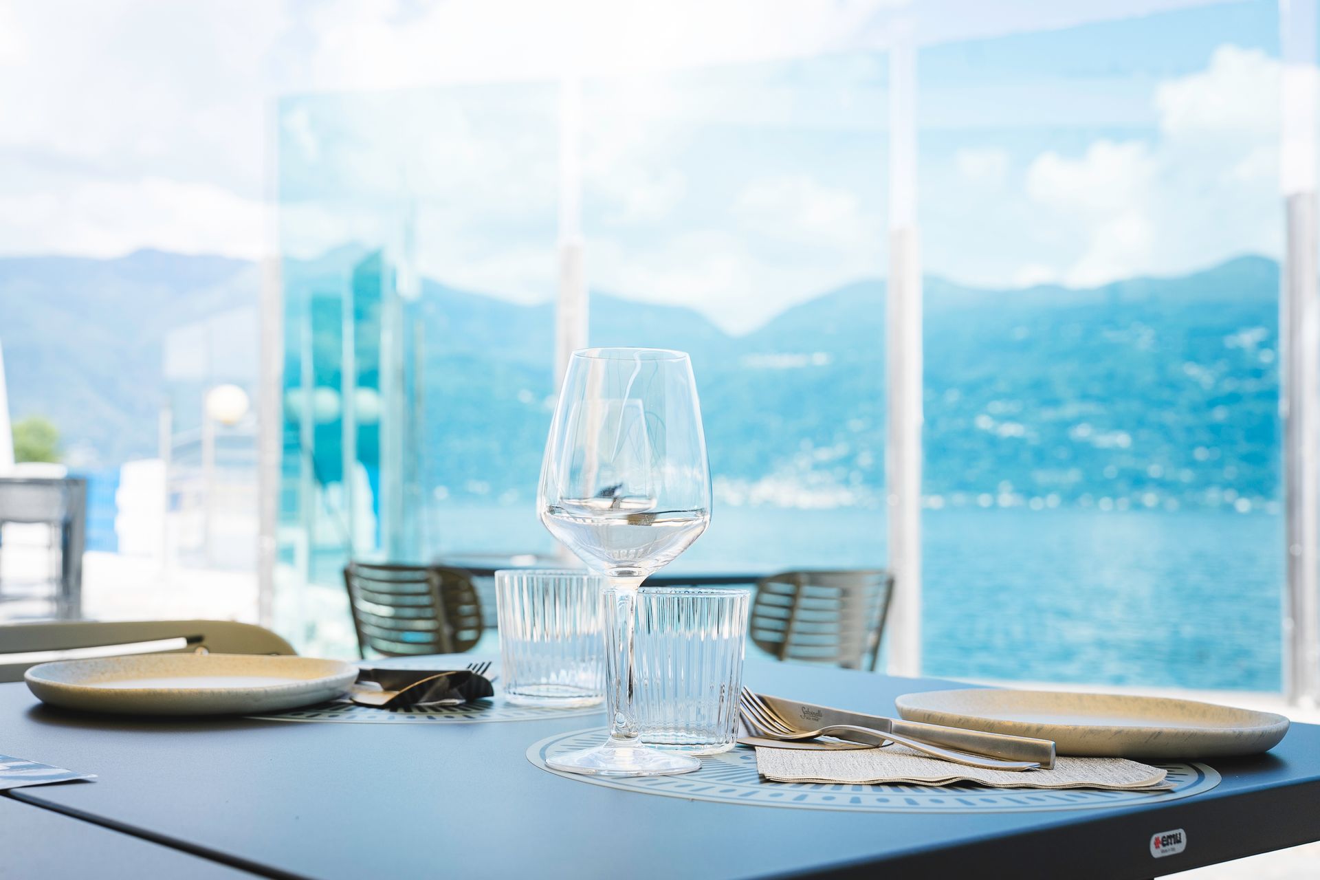 Tavolo da pranzo apparecchiato accanto a una luminosa finestra affacciata sul lago, con piatti, bicchieri e vista sulle montagne.