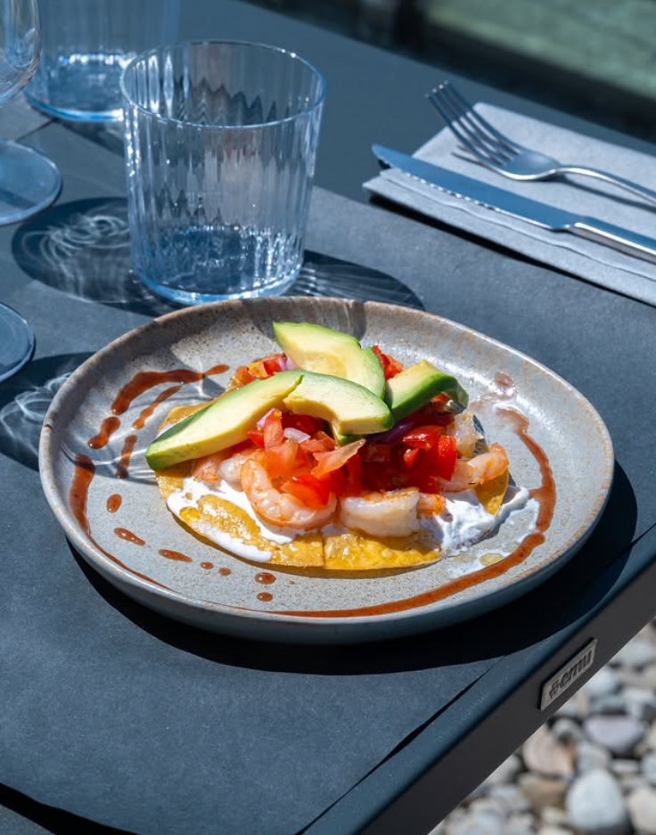 Colazione servita su un tavolo all'aperto con avocado in cima, bicchieri e posate a portata di mano.