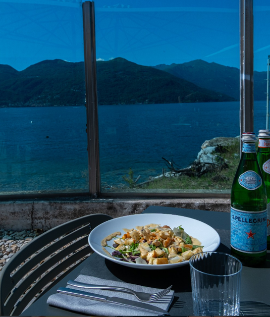 Un tavolo con la pasta in riva a un lago, con le montagne sullo sfondo e una bottiglia di acqua frizzante.