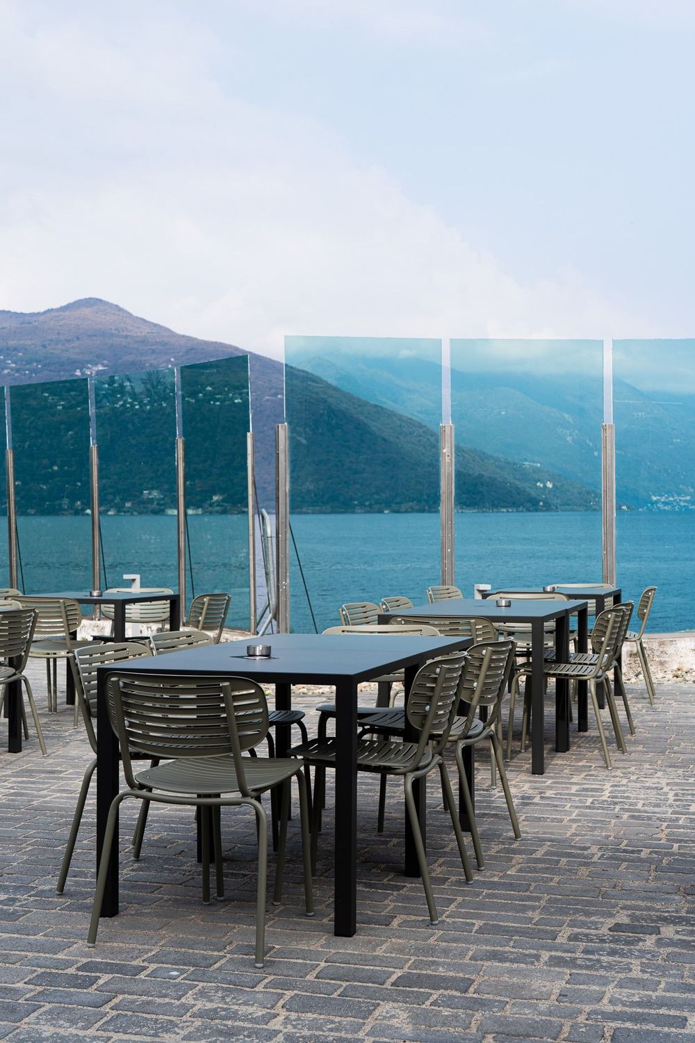 Tavolini e sedie da caffè all'aperto su una terrazza in pietra con vista su un lago azzurro e sulle montagne.