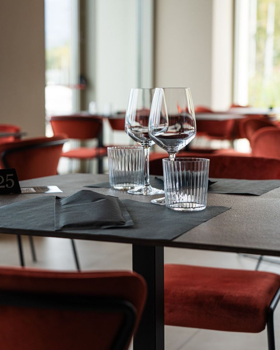 Tavolo da ristorante apparecchiato con bicchieri da vino, bicchieri d'acqua, tovaglioli scuri e sedie rosse sullo sfondo.