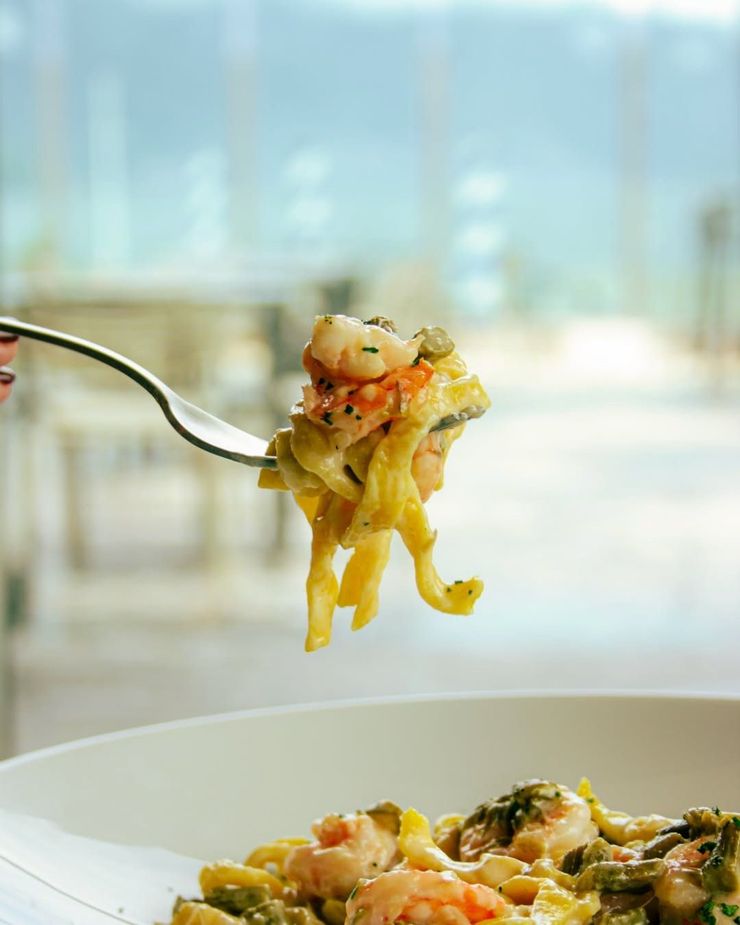 Una forchetta solleva la pasta con le verdure da una ciotola bianca in un ambiente luminoso e sfocato adibito a sala da pranzo.