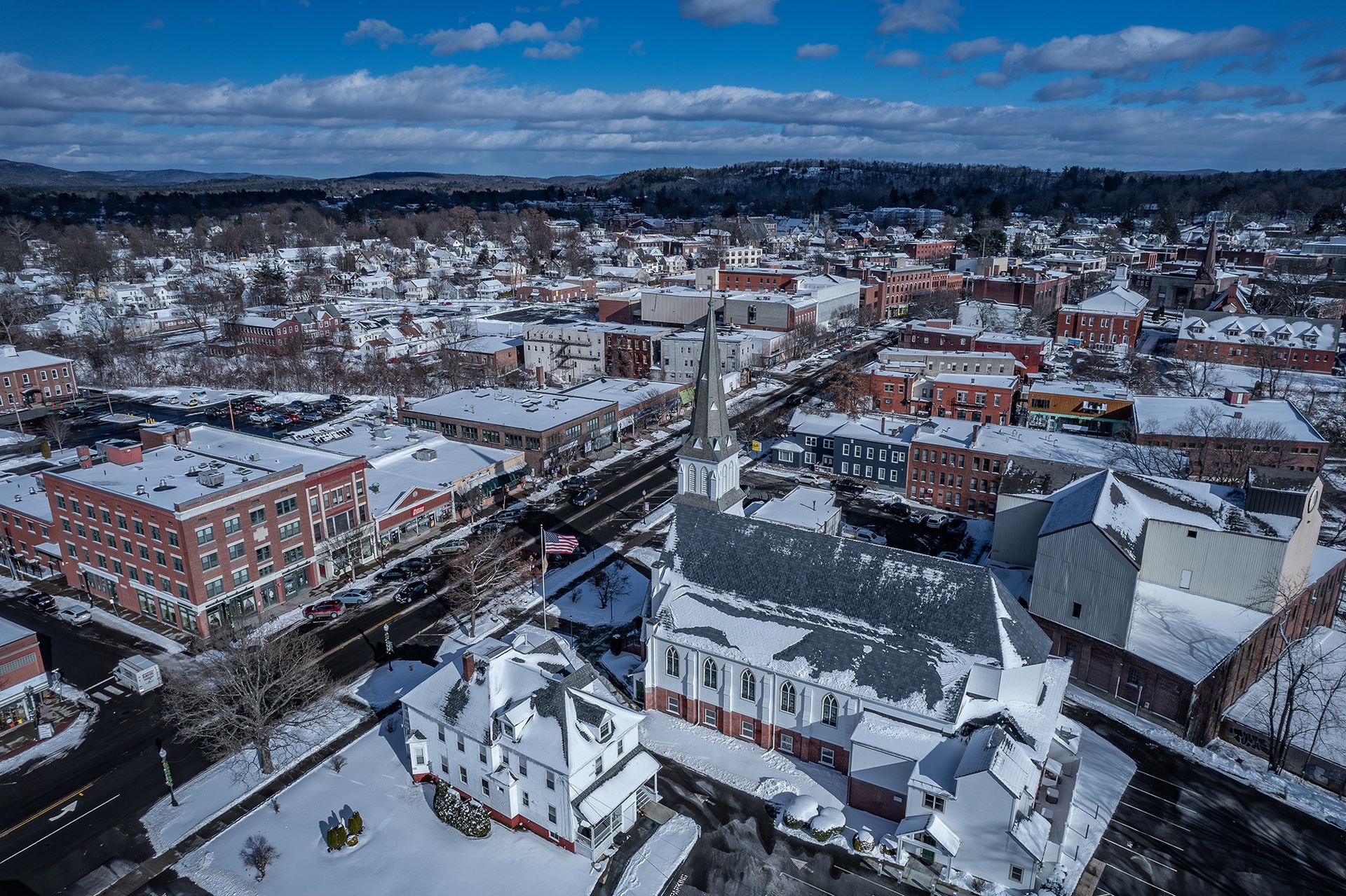 Snowy aerial view of a town with a church tower, rooftops, and streets under a cloudy blue sky