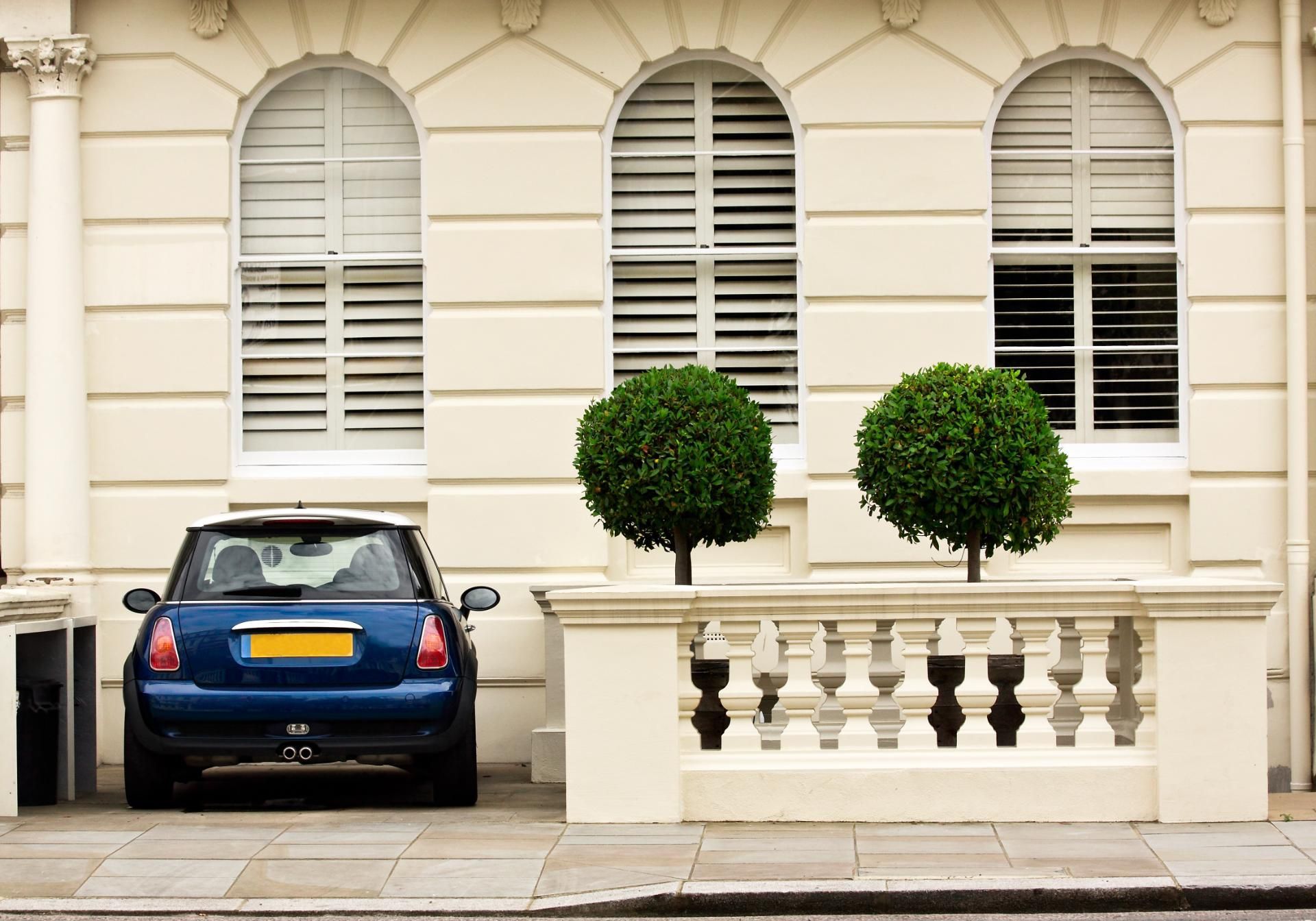 Blue car parked beside white townhouse with arched windows and trimmed topiary trees.