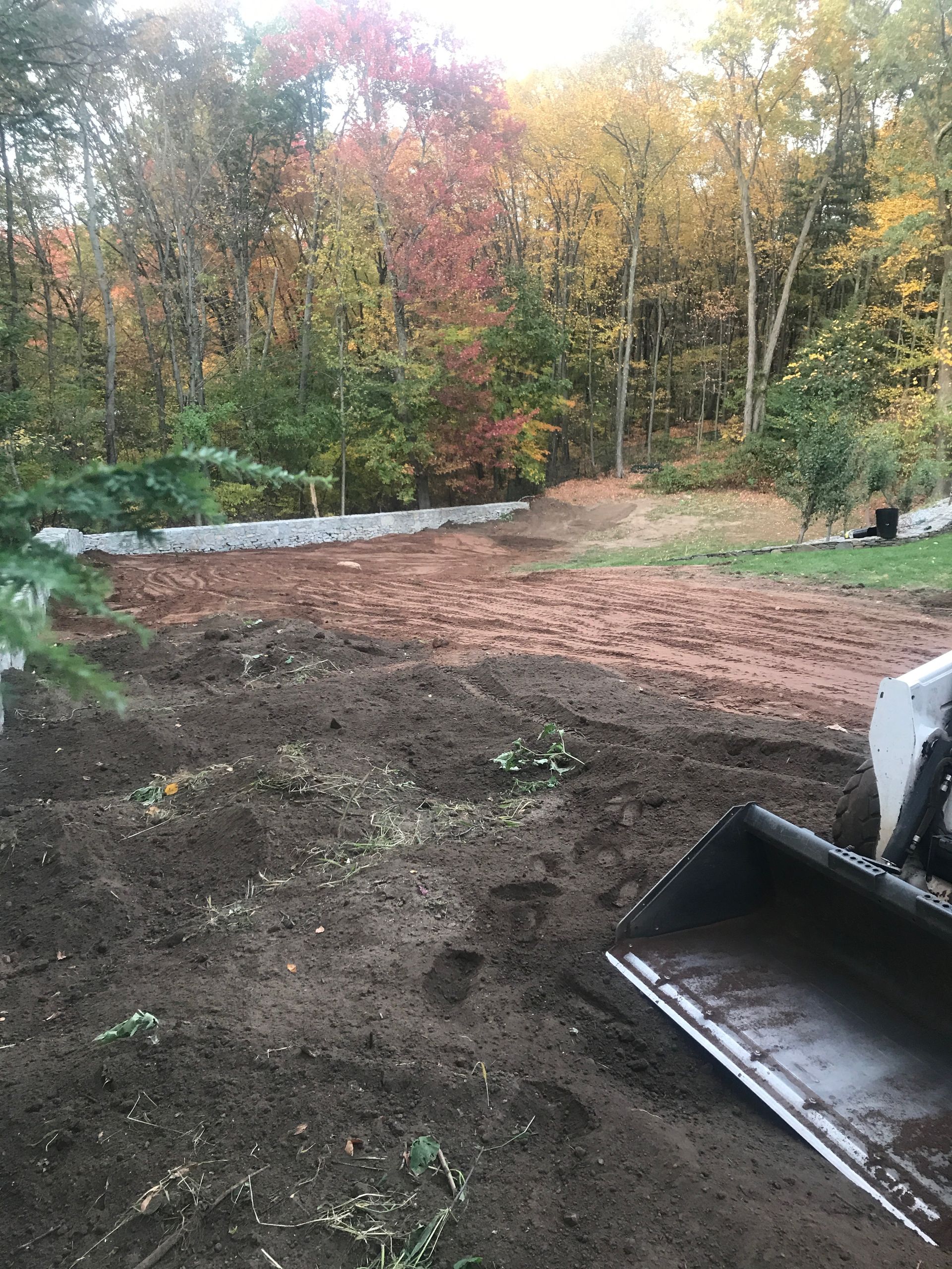 Excavator on a dirt lot, preparing a yard for landscaping. Fall foliage in the background.