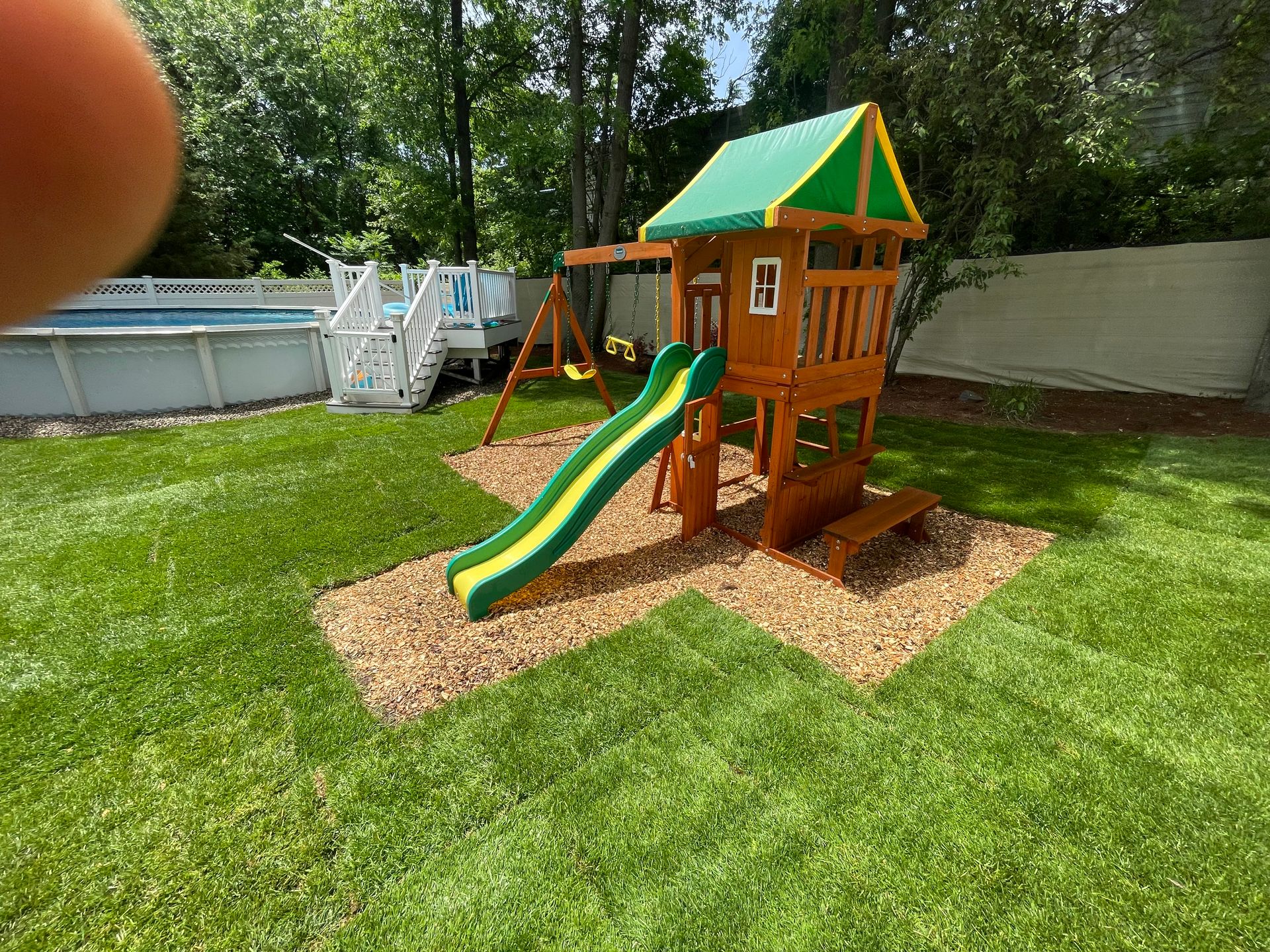 Wooden playset with slide and swing set in a backyard with green grass, near a swimming pool.