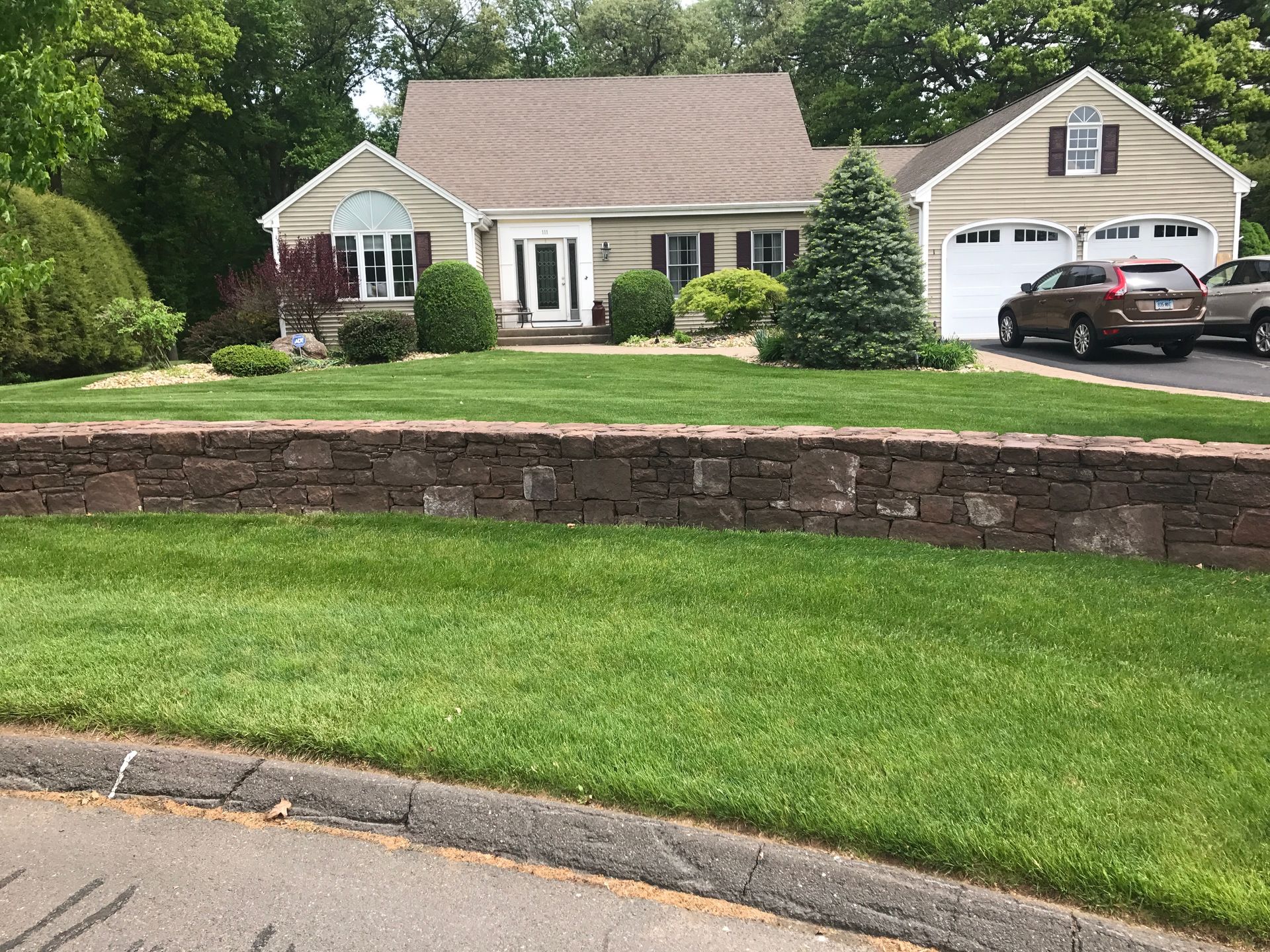 Curb-side view of a beige house with a stone retaining wall and green lawn. Cars parked in driveway.