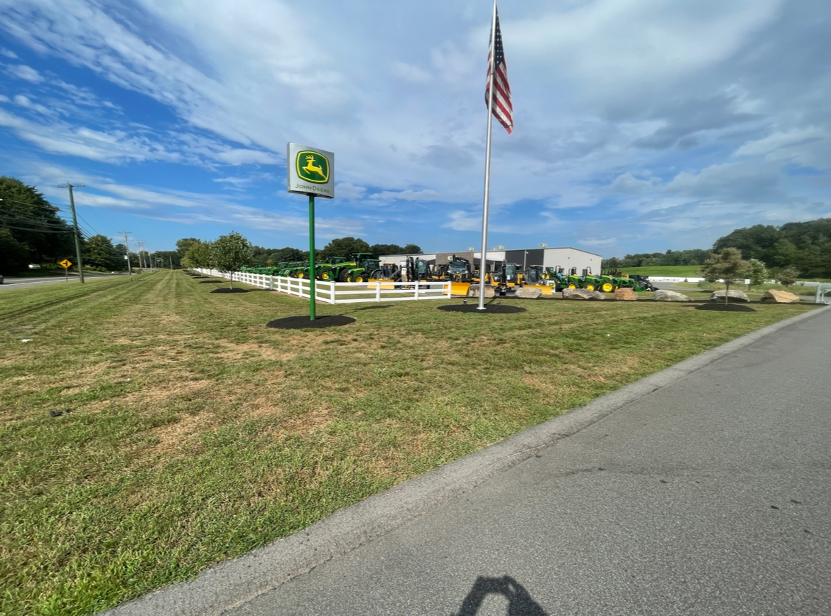 John Deere dealership with a sign, American flag, and equipment displayed on a sunny day.