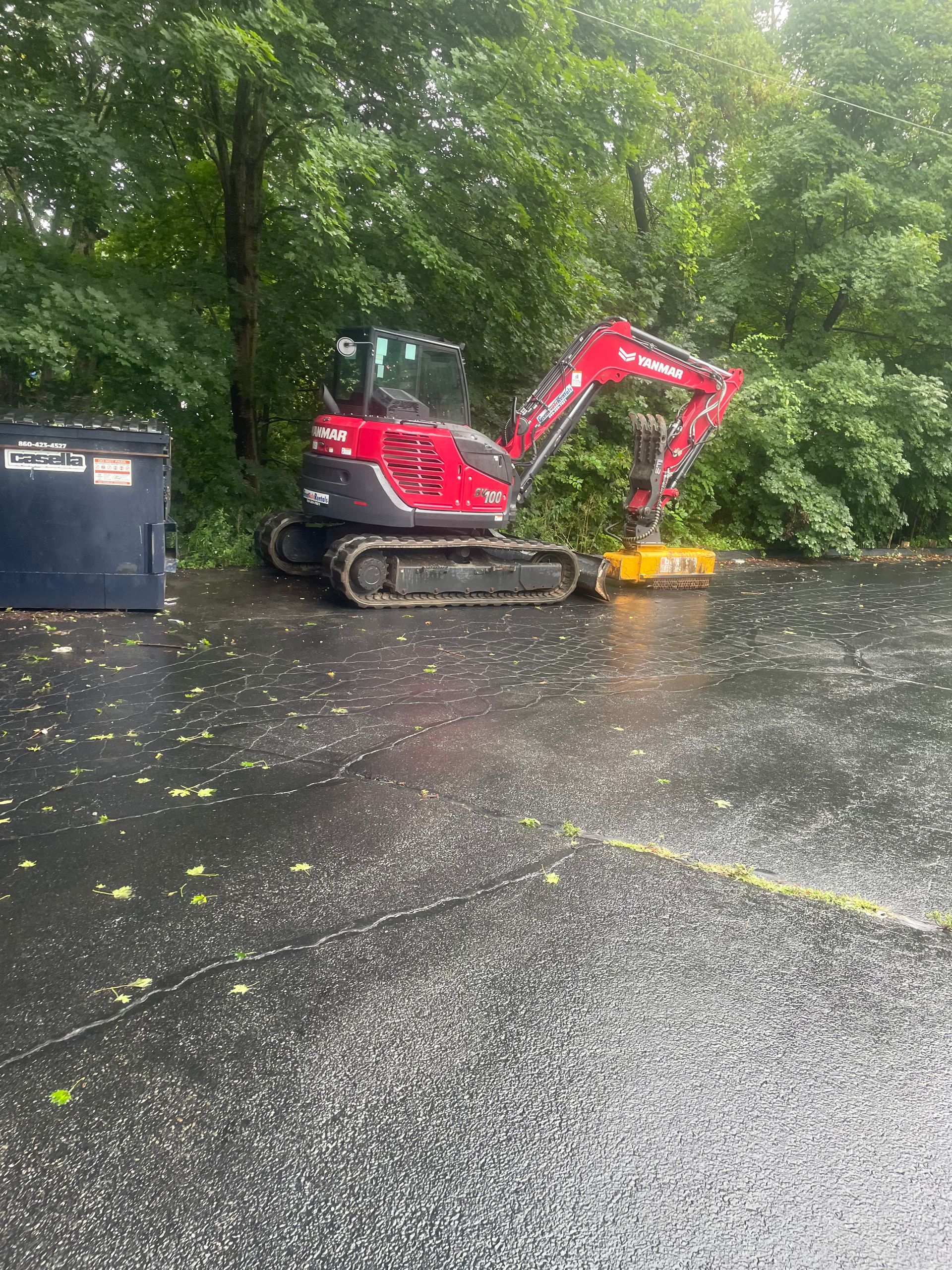Red excavator with a yellow attachment in a parking lot, near a dumpster and trees.