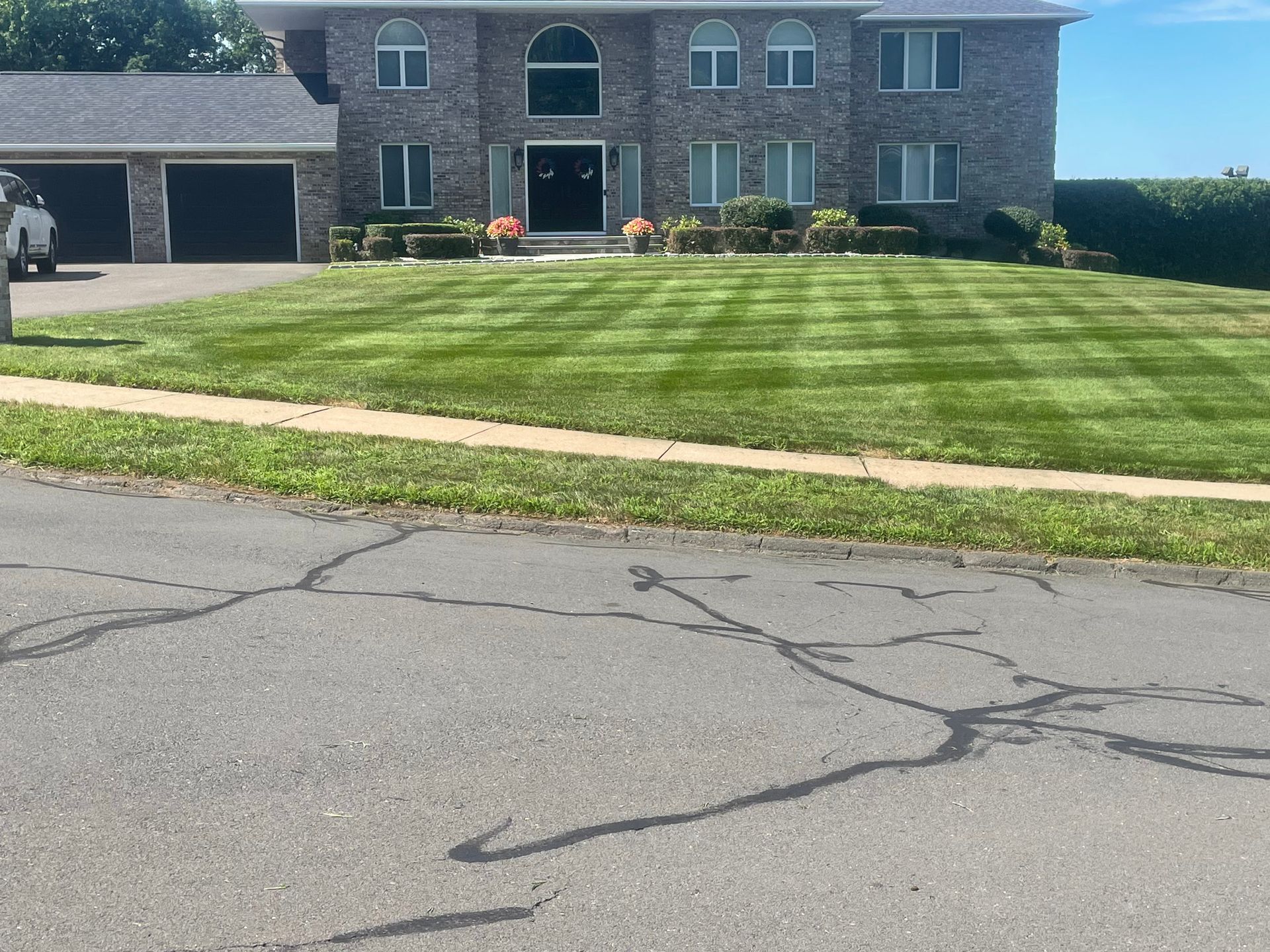 Two-story brick house with striped lawn, asphalt street in foreground, and blue sky.