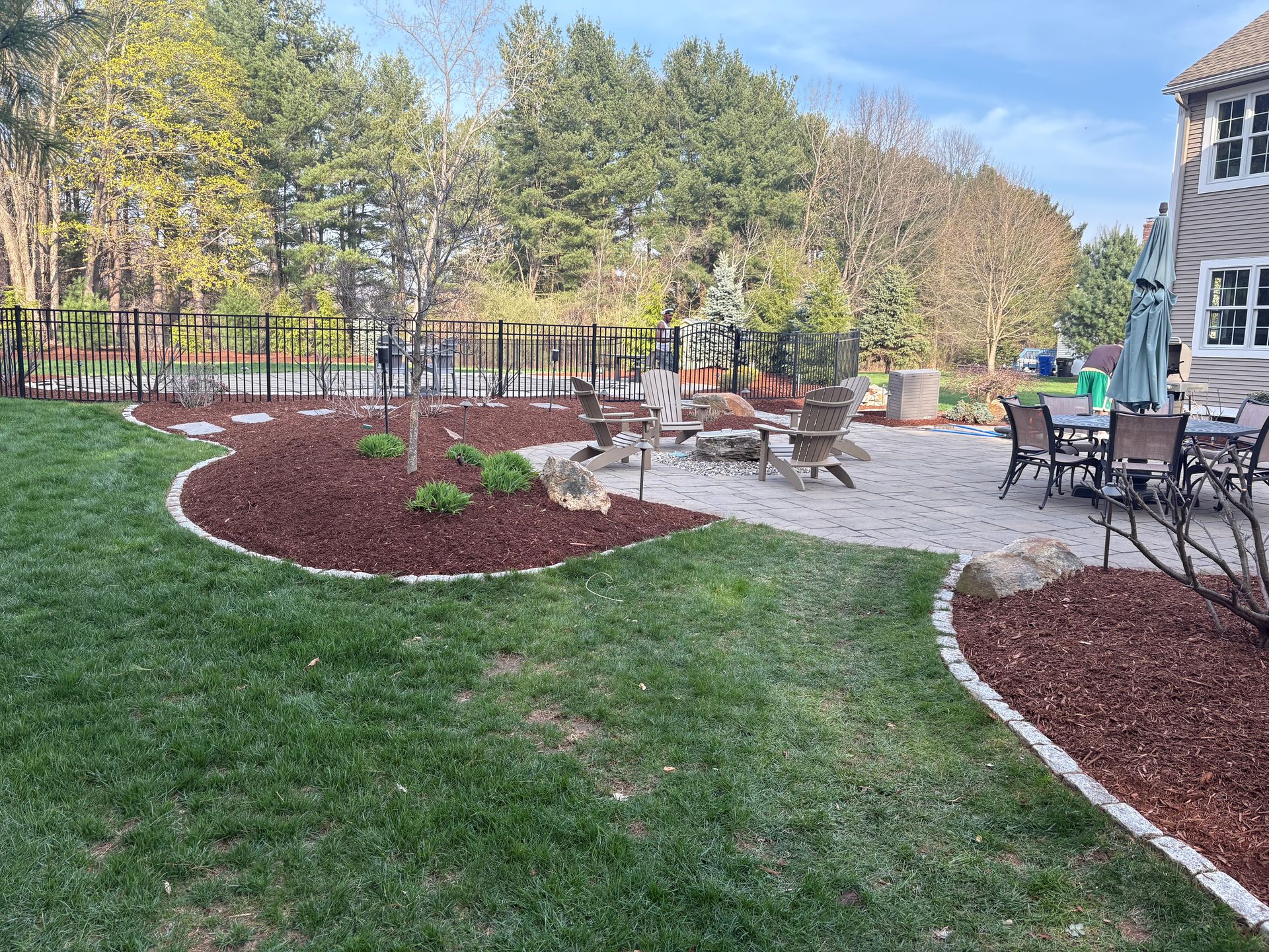 Backyard with patio, fire pit, dining set, and landscaped beds. Green grass surrounds.