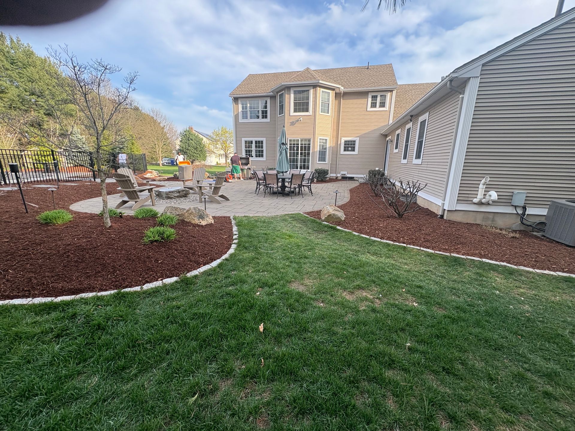 Backyard with patio, lawn, and house. Mulched flowerbeds border a circular stone patio with outdoor furniture.