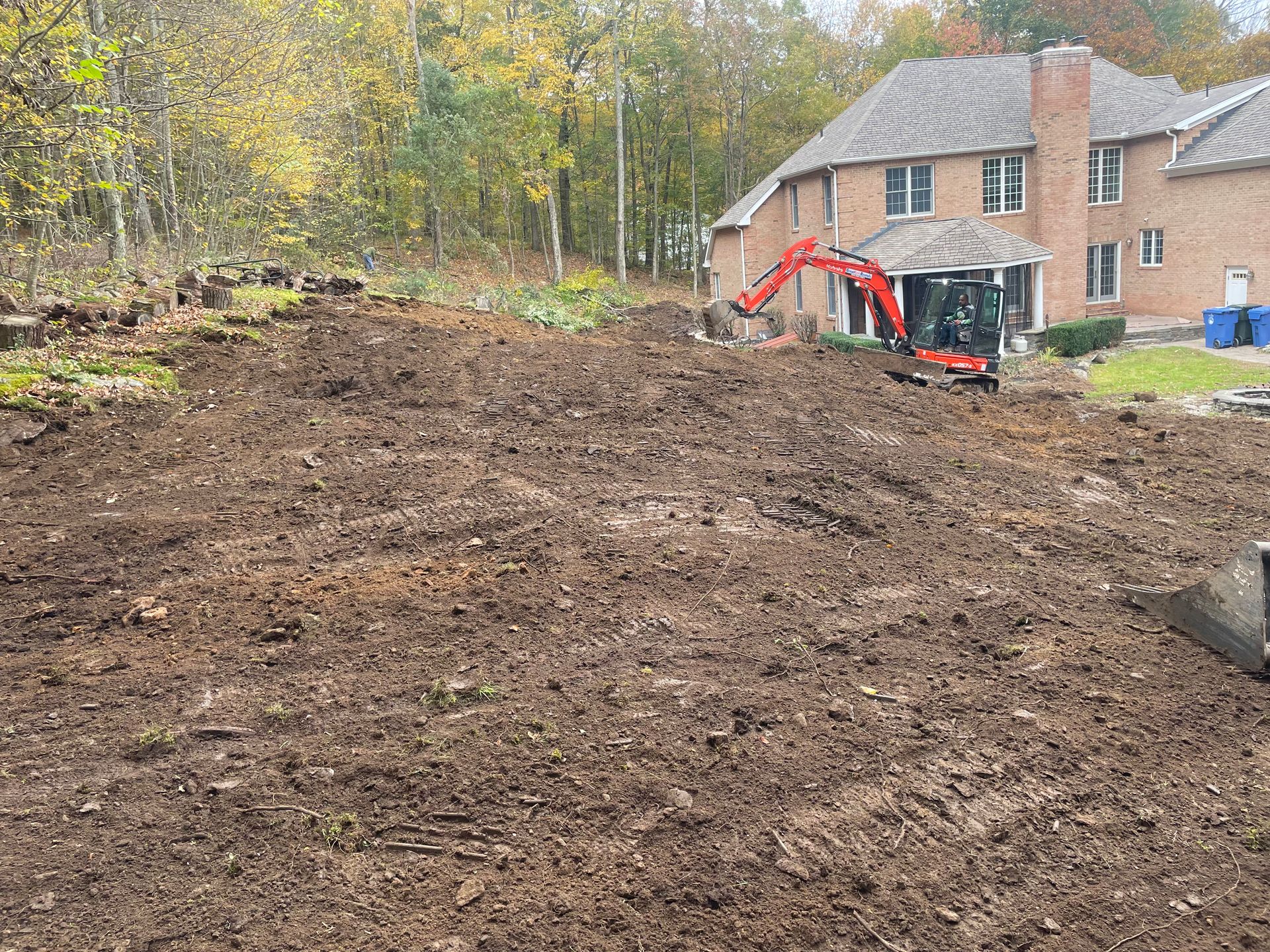 An excavator working on a muddy, cleared yard in front of a large house; trees in the background.