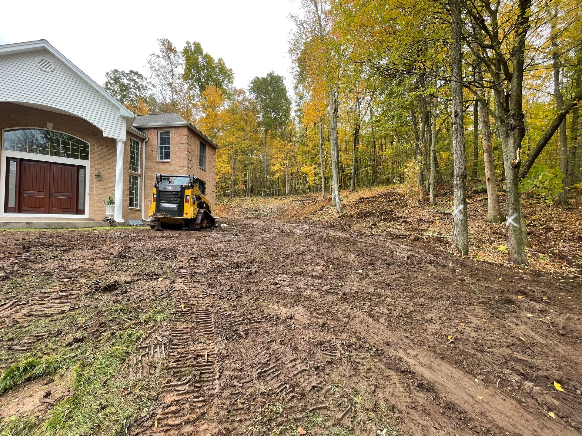 A skid steer on muddy ground next to a house with trees and fall foliage.