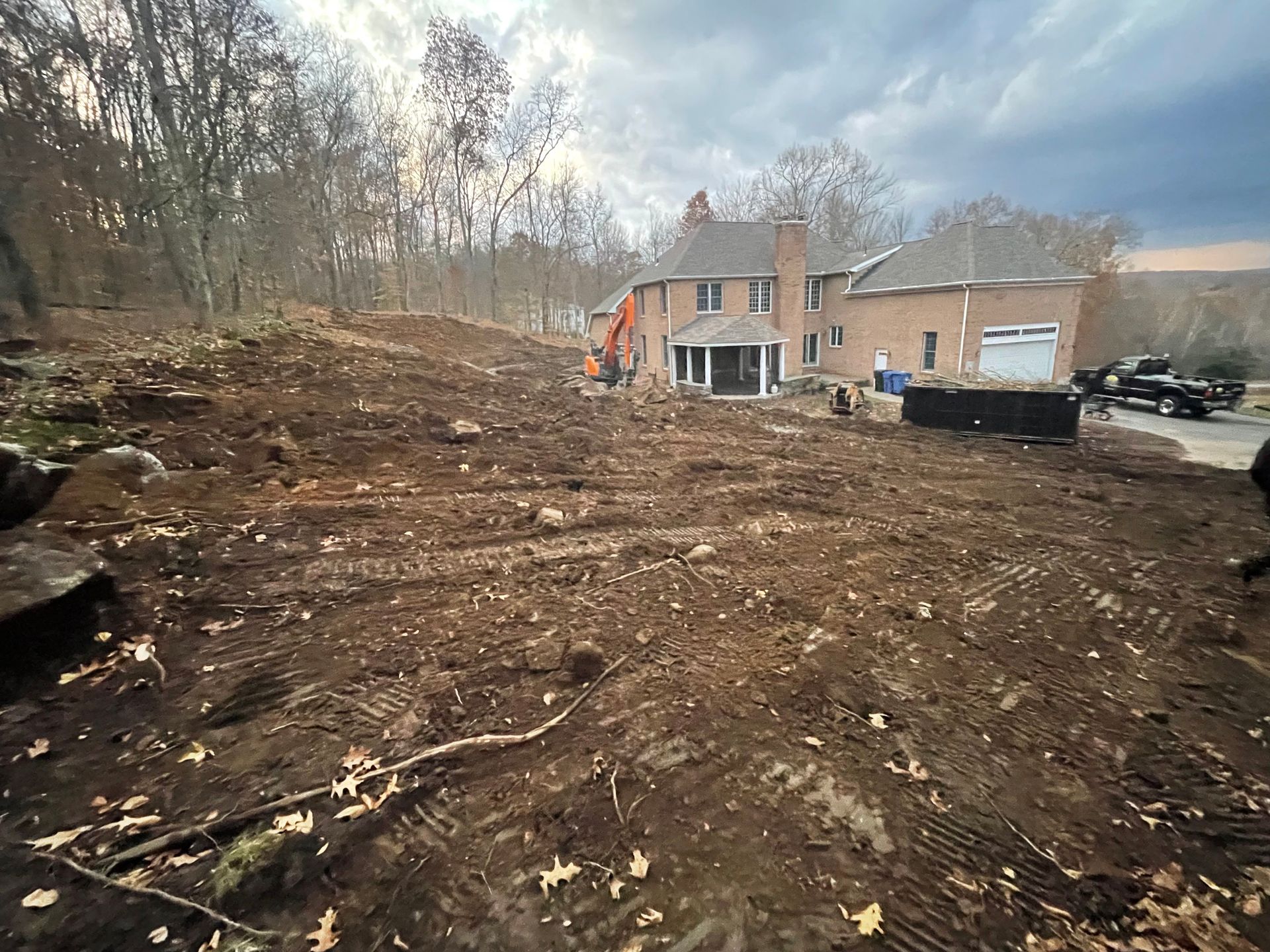 Cleared lot in front of a brick house; dirt and rocks in foreground, excavator visible, trees in background.