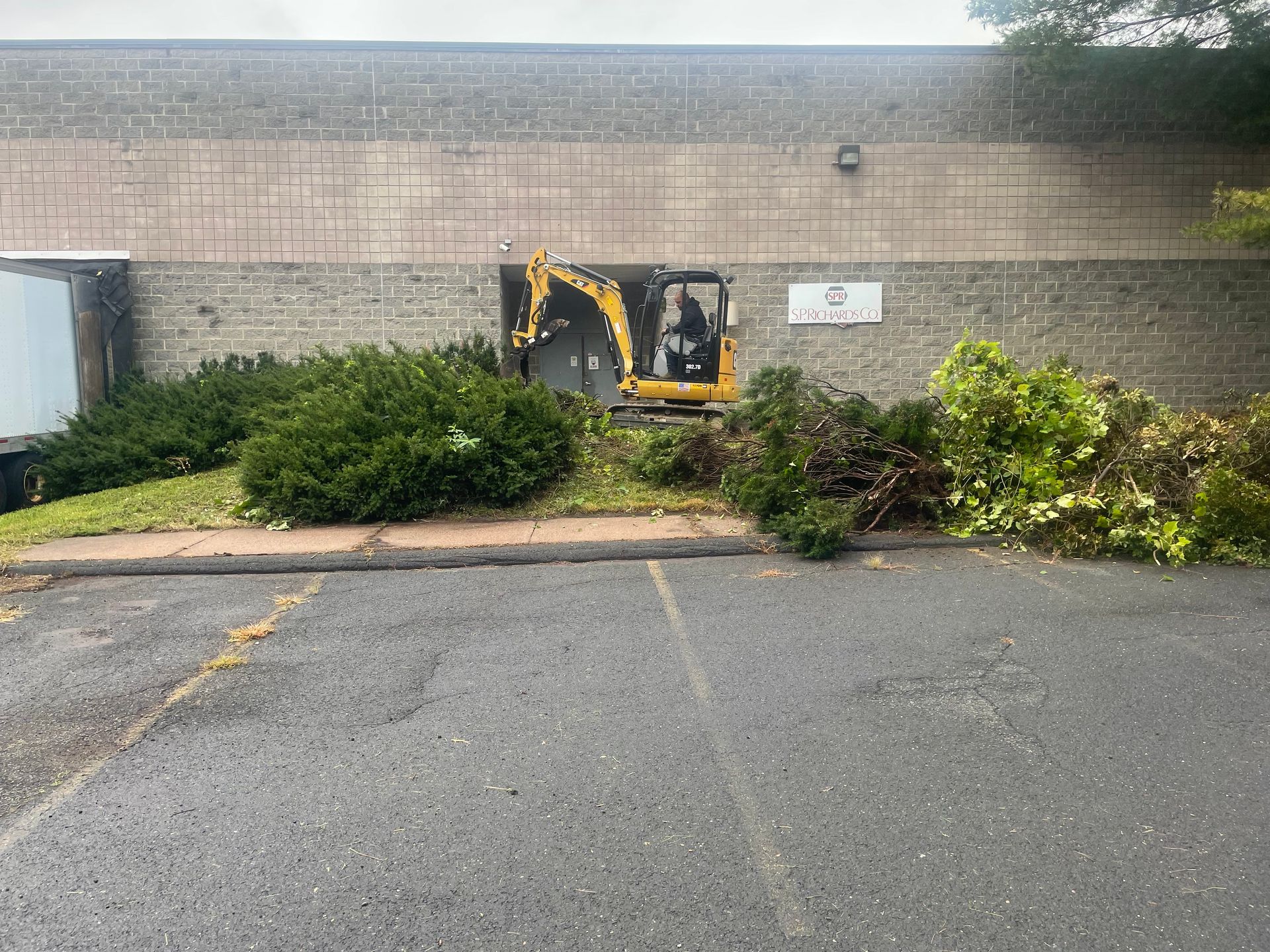 Mini-excavator removing vegetation in front of a brick building. Debris piled on ground; overcast.