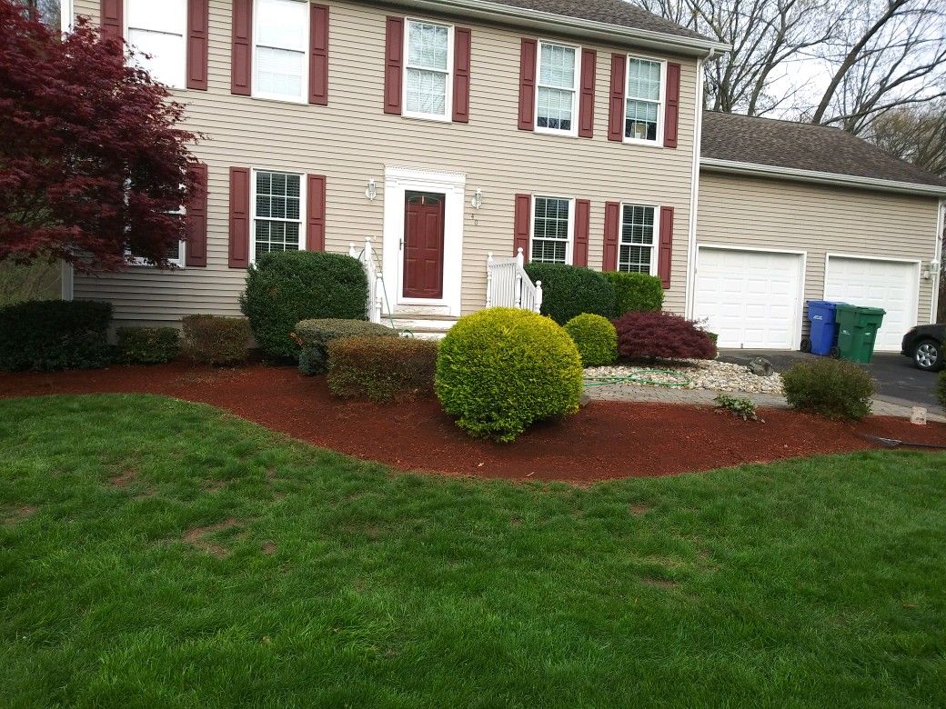 Beige house with red shutters, brown mulch beds with green shrubs, green grass lawn, two car garage.