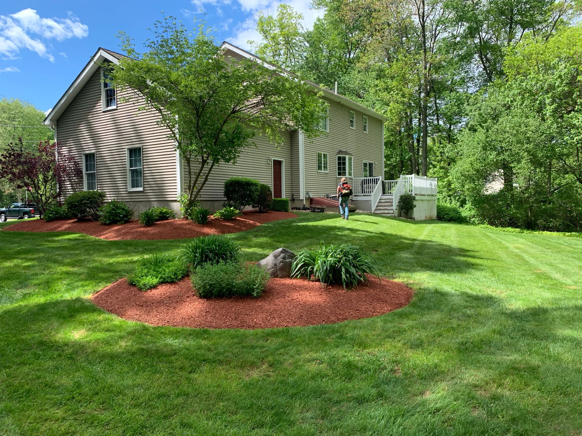 Two-story house with green lawn and red mulch beds. A person stands on the porch. Trees surround the house.
