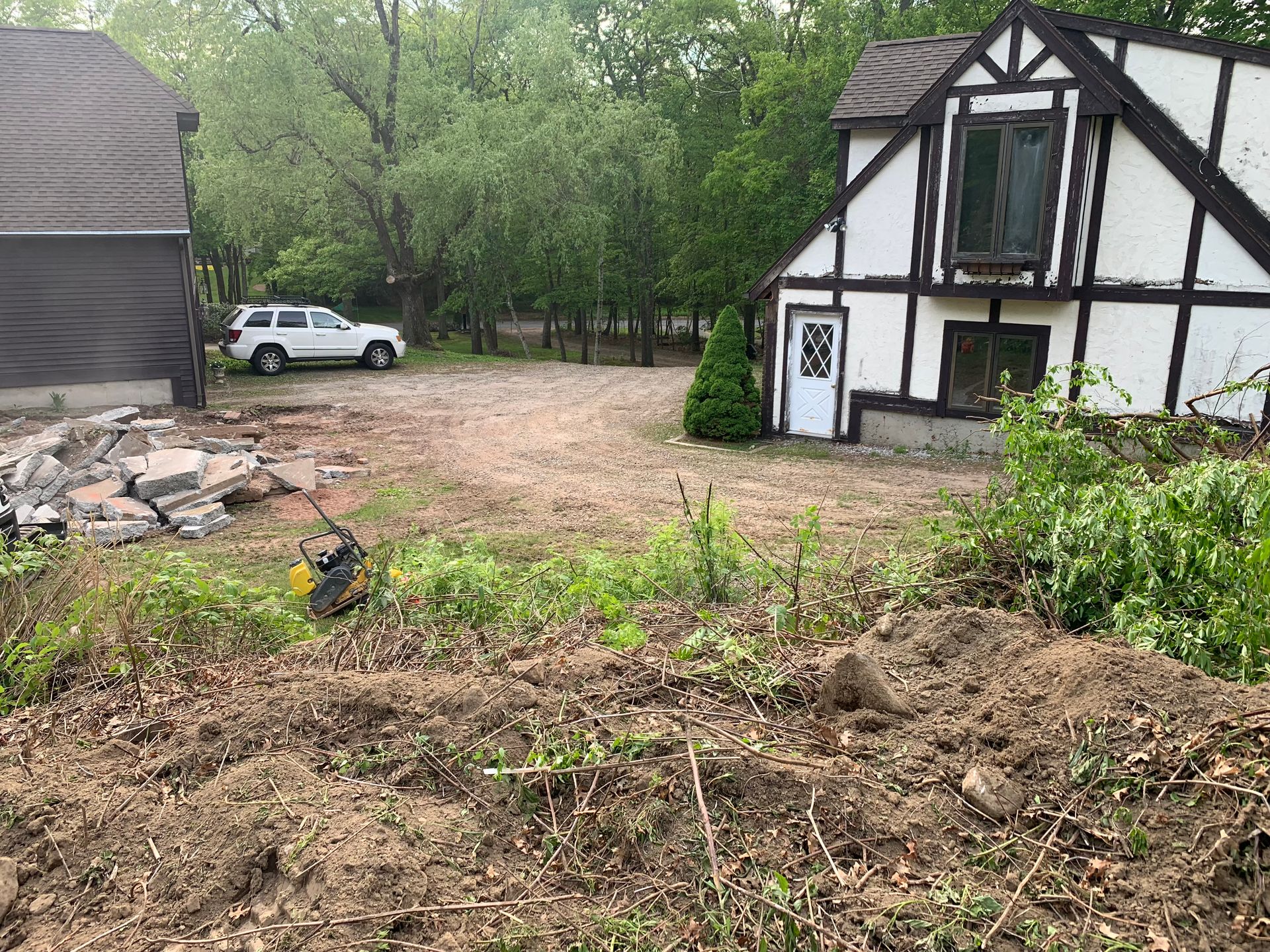 Cleared lot with two buildings: Tudor-style house and a building with a dark roof. White SUV parked in the middle.