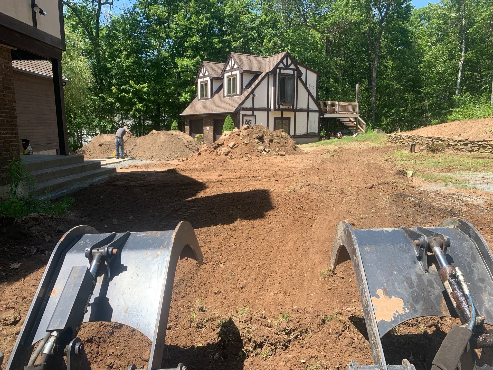 Excavator buckets in foreground; dirt work in progress near Tudor-style house and steps. Man shoveling in background.