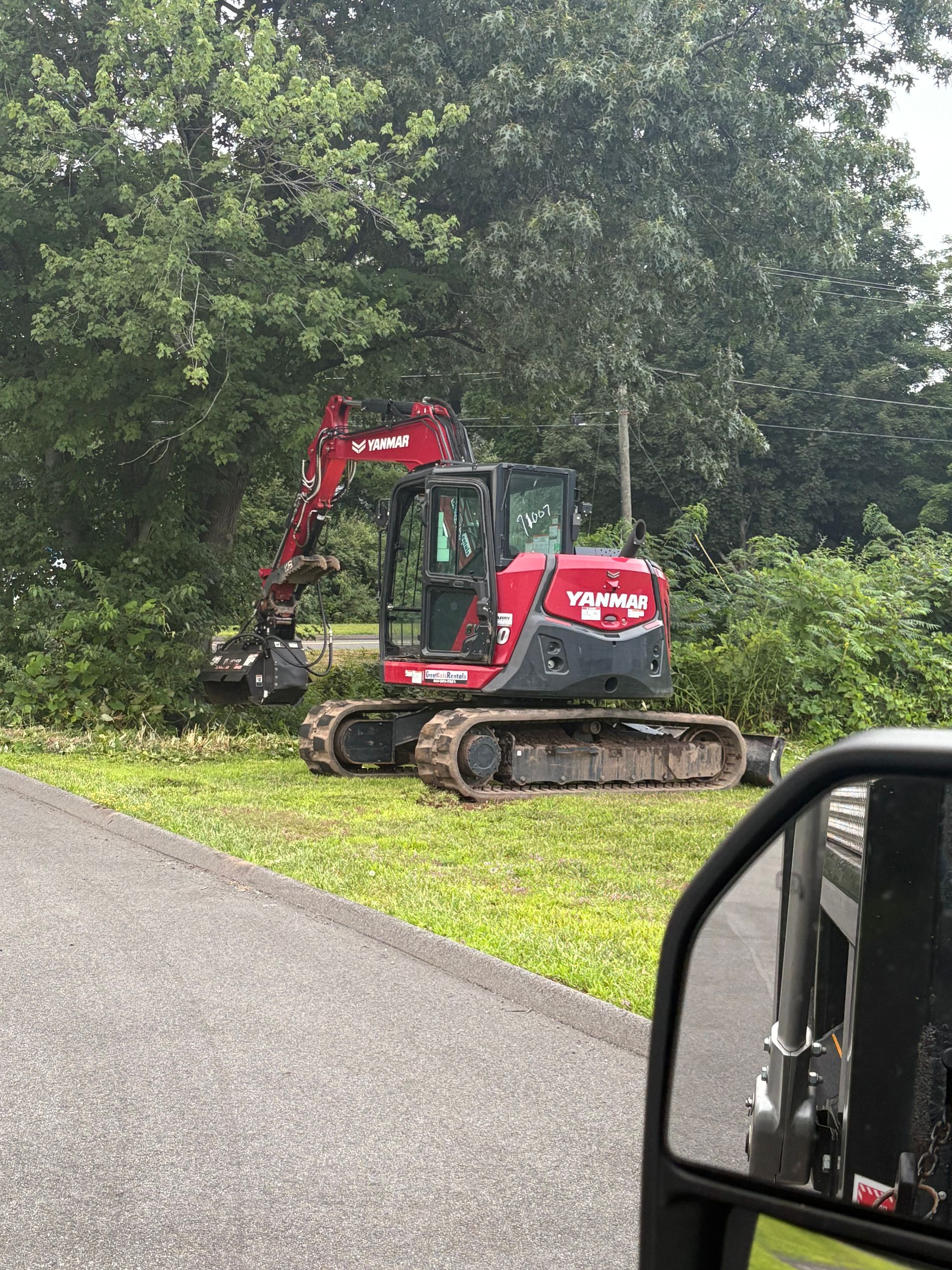 Red and gray excavator trimming bushes beside a road, seen from a vehicle's side mirror.