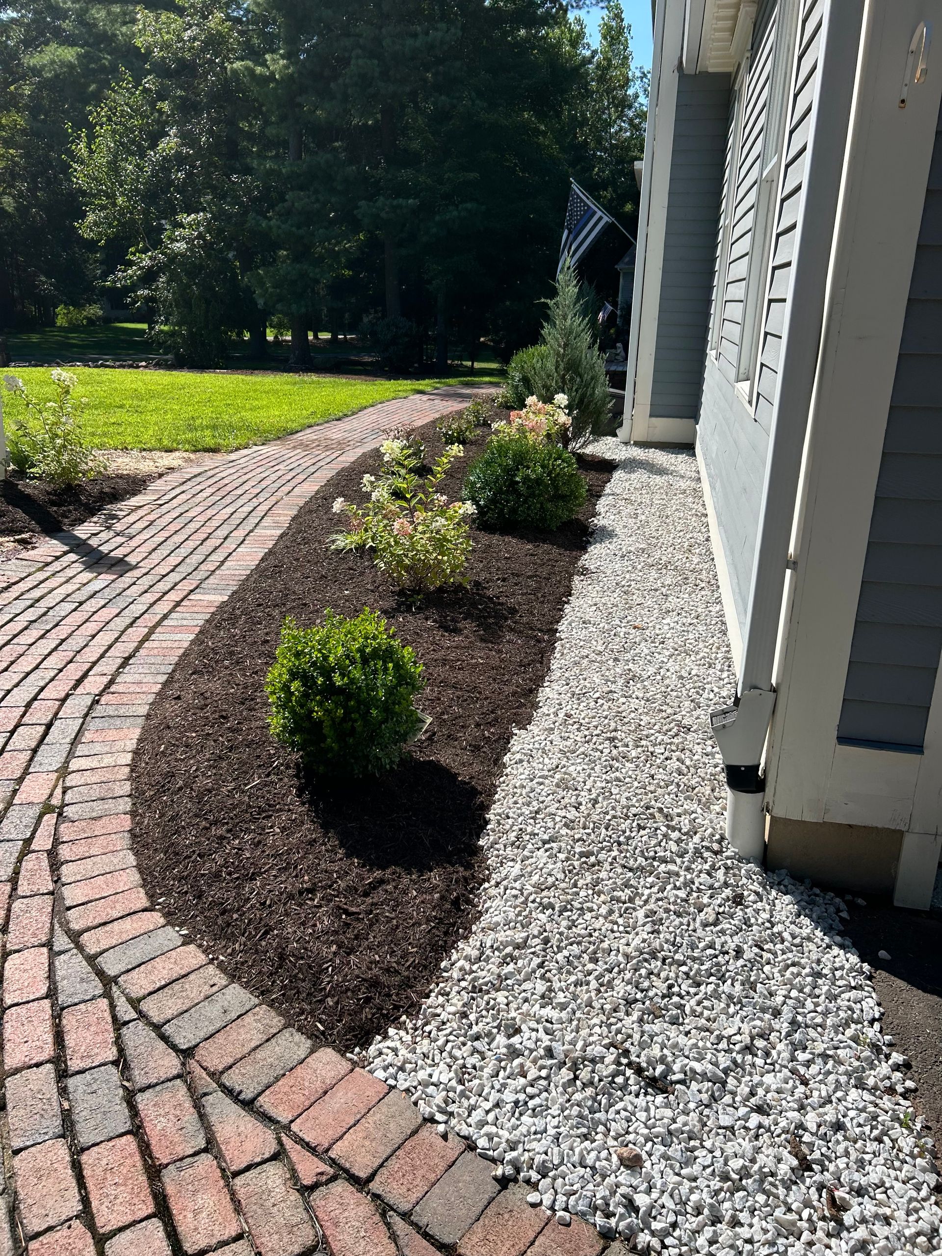 Landscaped flower bed with brick walkway, mulch, gravel, and house with gray siding.