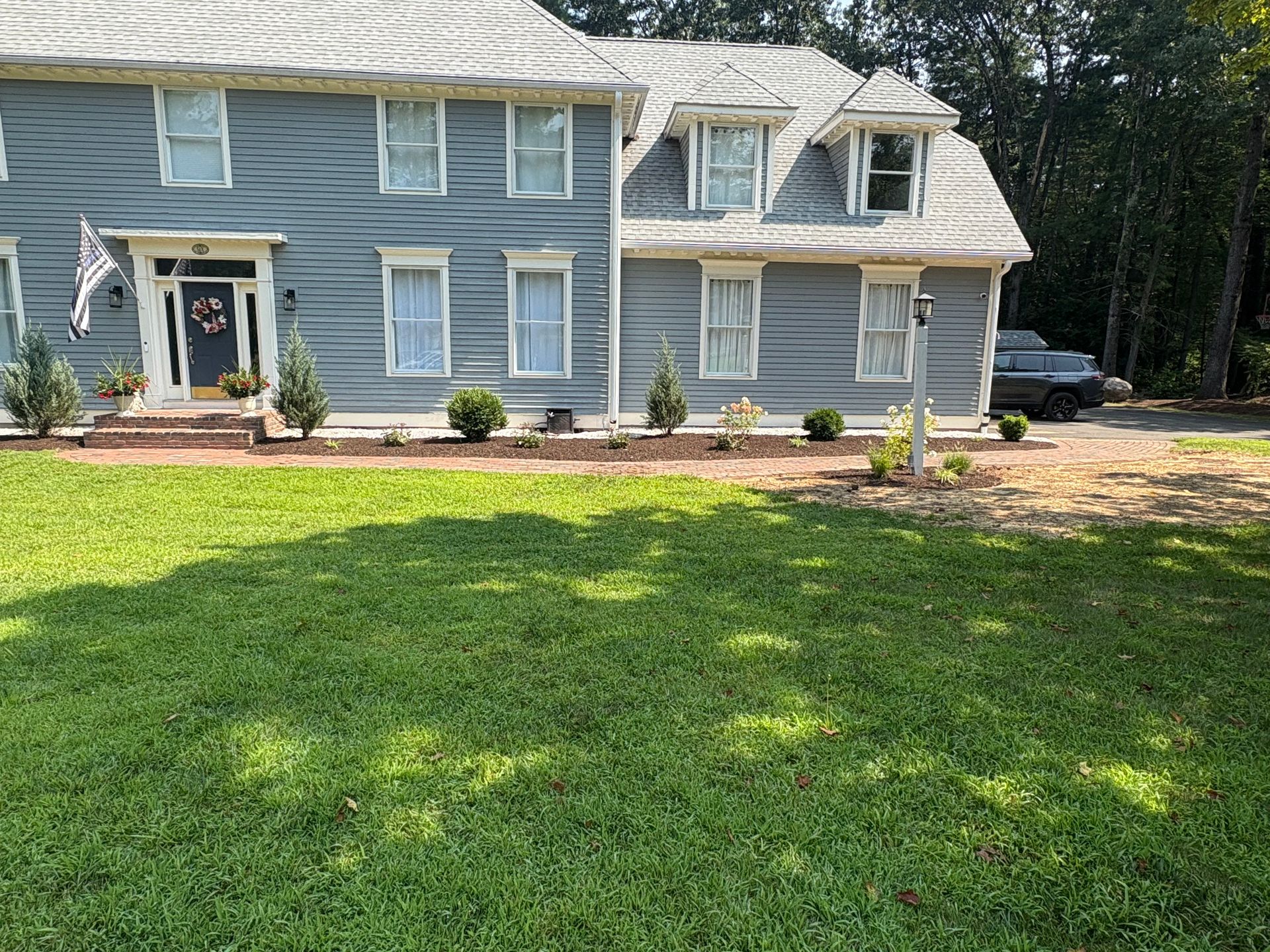 Two-story blue house with white trim. Green lawn and landscaped front yard with a car parked nearby.