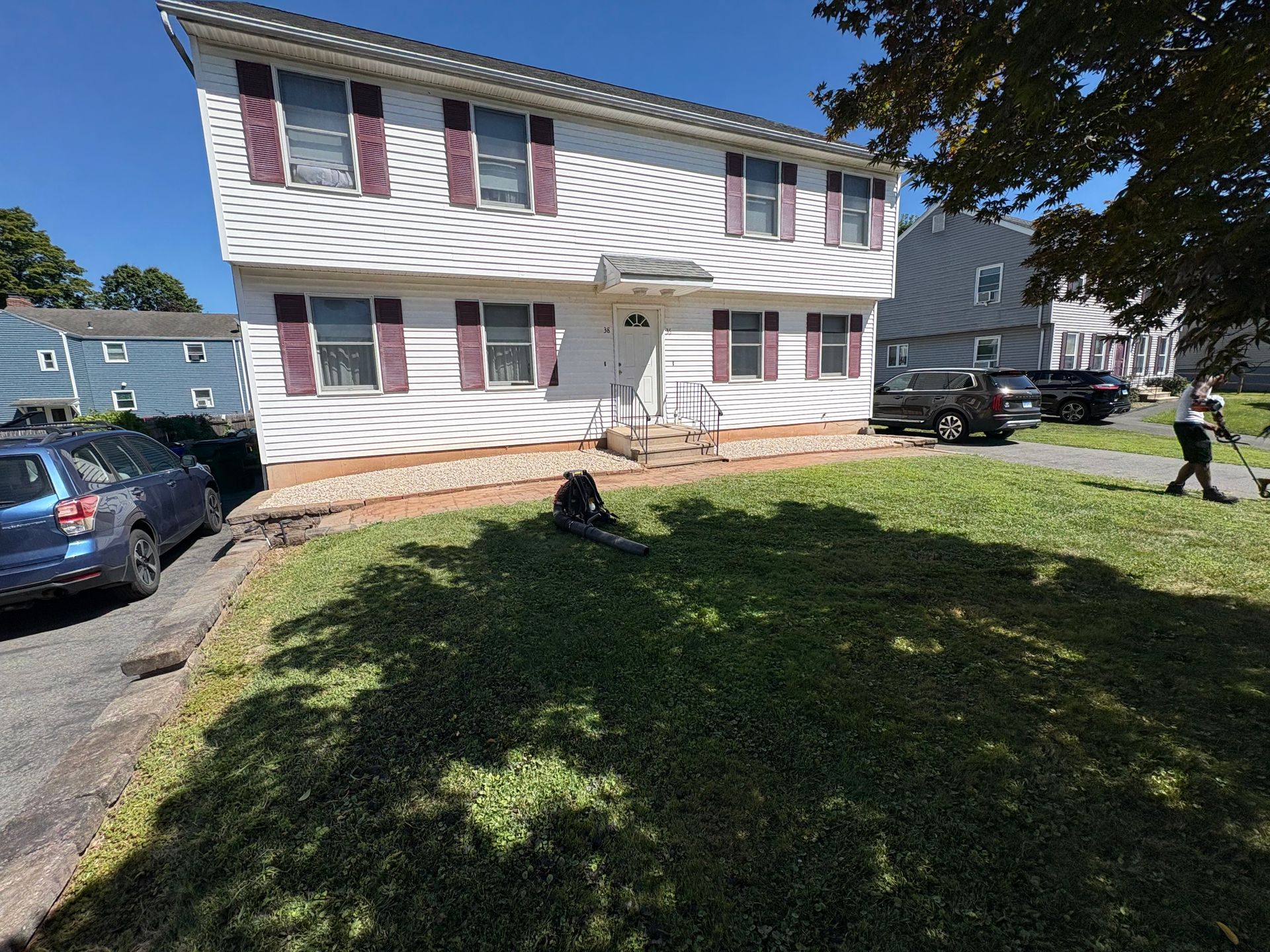 Two-story white house with maroon shutters and a person mowing the lawn on a sunny day.