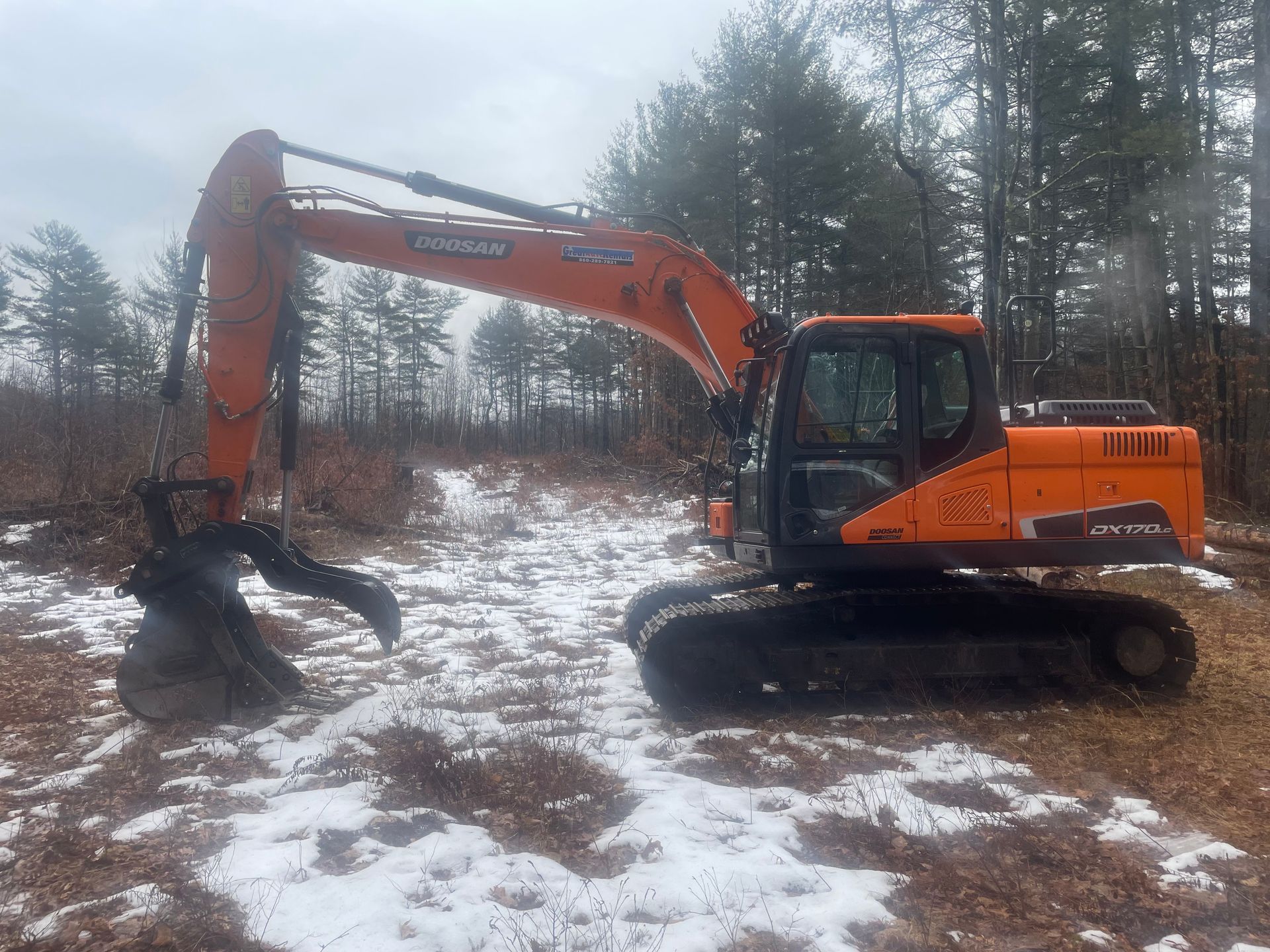 Orange Doosan excavator with grapple attachment in a snowy, wooded area.